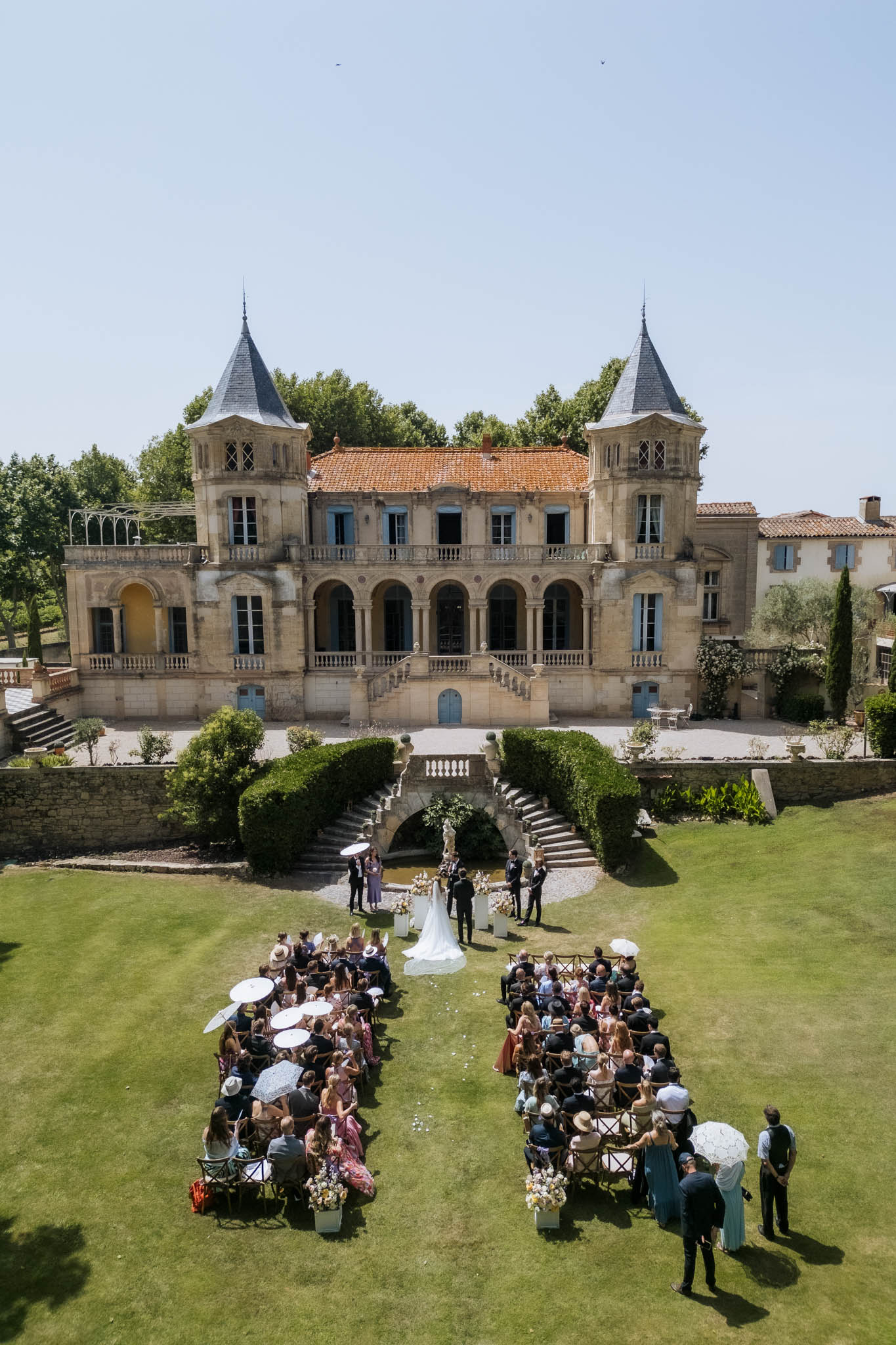 Aerial view of outdoor ceremony at turreted chateau with white floral arch, urns, and guests with parasols