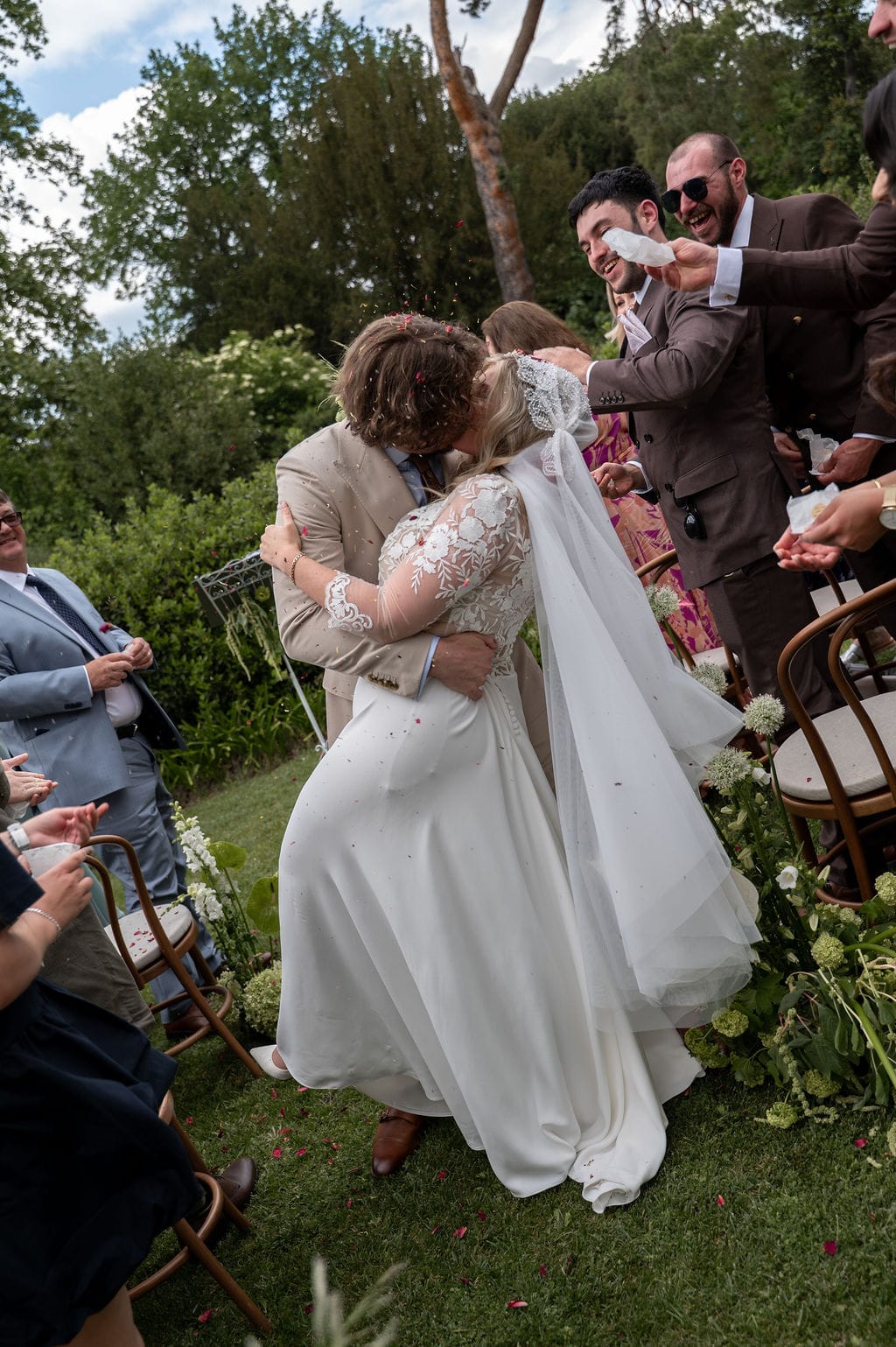 Couple kissing during first kiss with red confetti toss guests on bentwood chairs and white aisle florals
