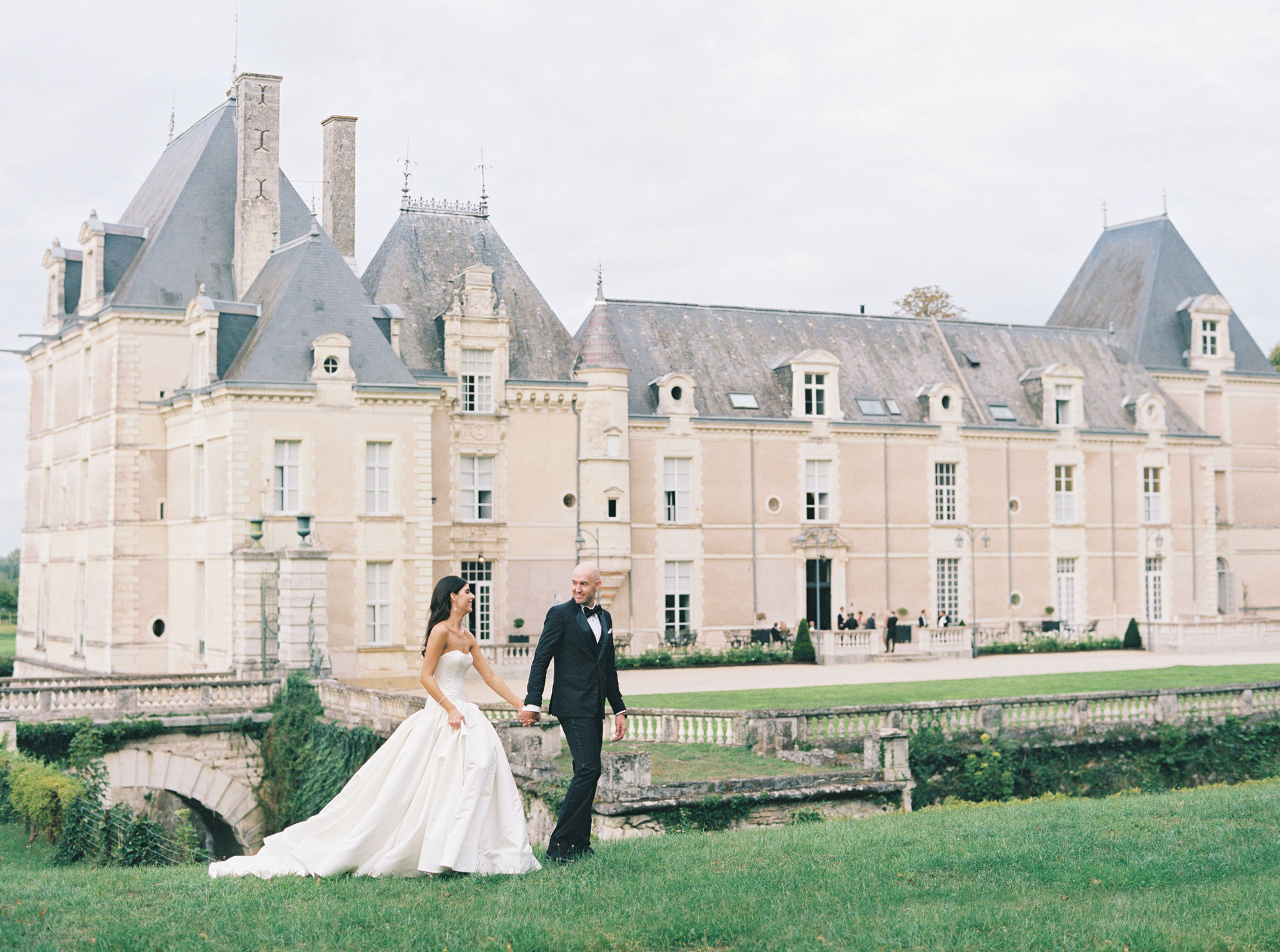 Couple walking hand-in-hand on lawn before classical chateau with slate towers and stone balustrade