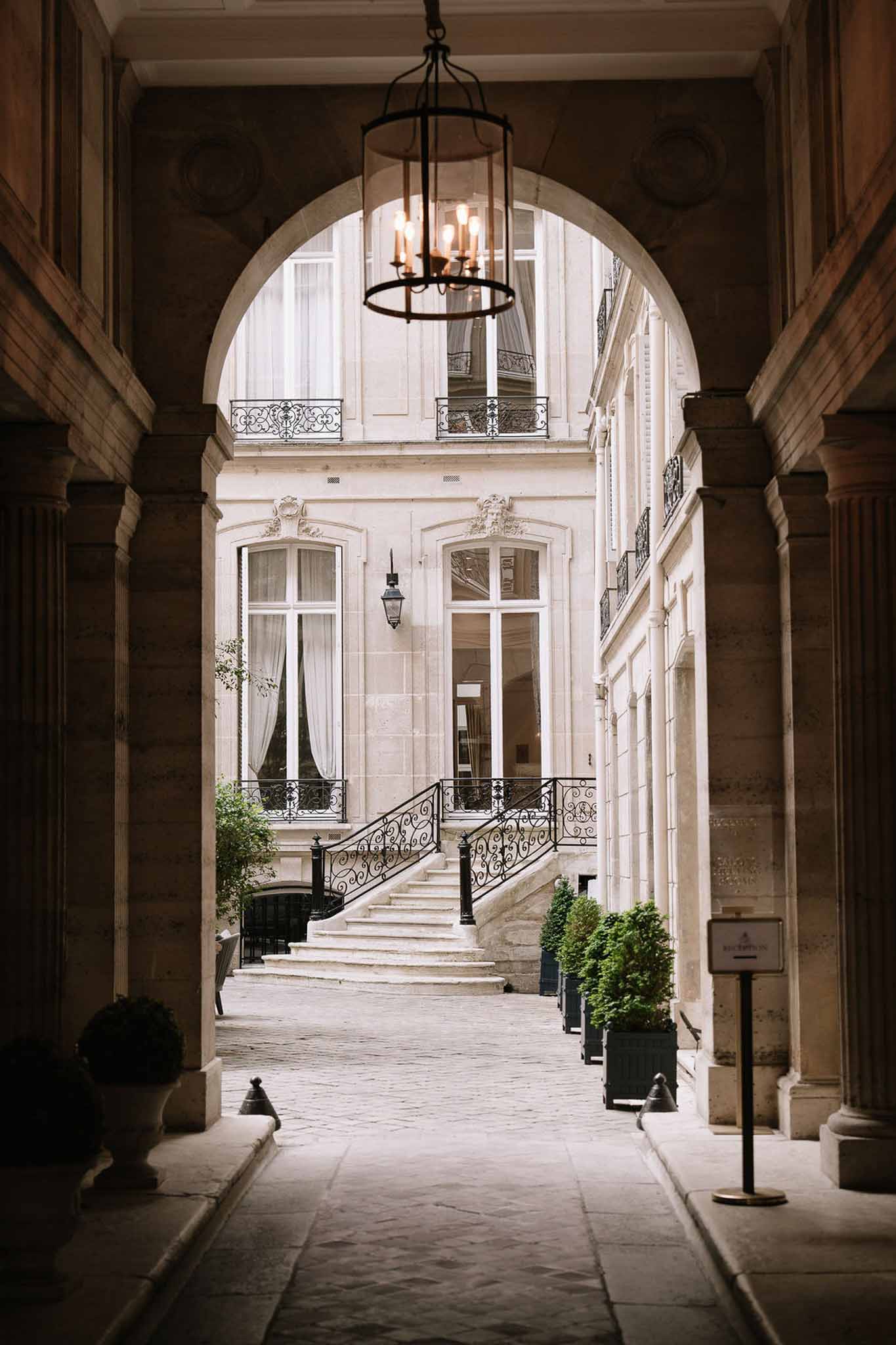 View through stone arch into Haussmann-style Parisian courtyard with grand staircase, wrought-iron railings, and boxwood t...