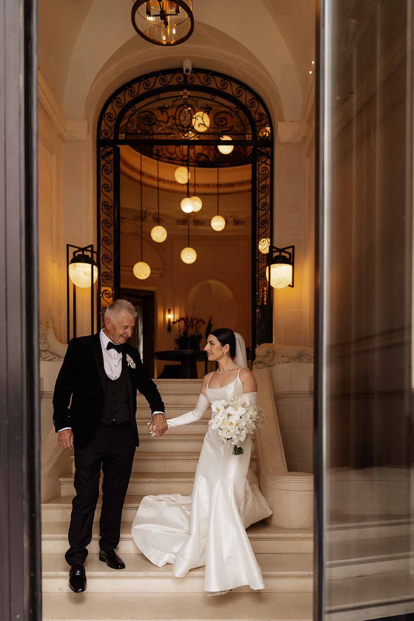 Bride and father descend marble staircase through ornate iron gate, bride in off-shoulder gown with orchid bouquet