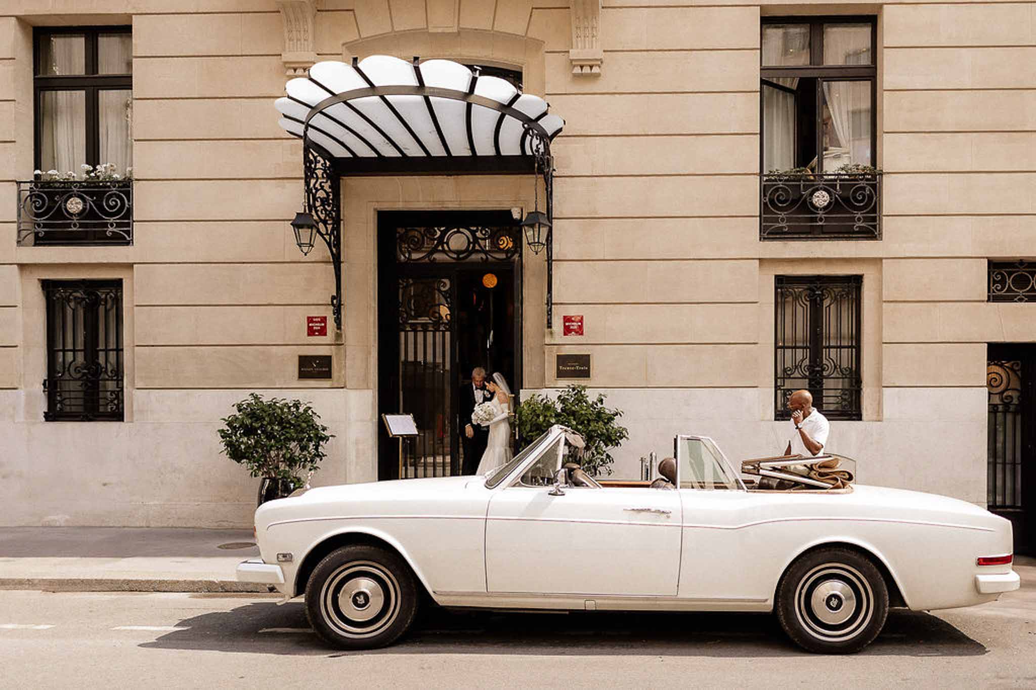 Couple exiting Haussmann building beside vintage white Rolls-Royce, bride in white gown with bouquet
