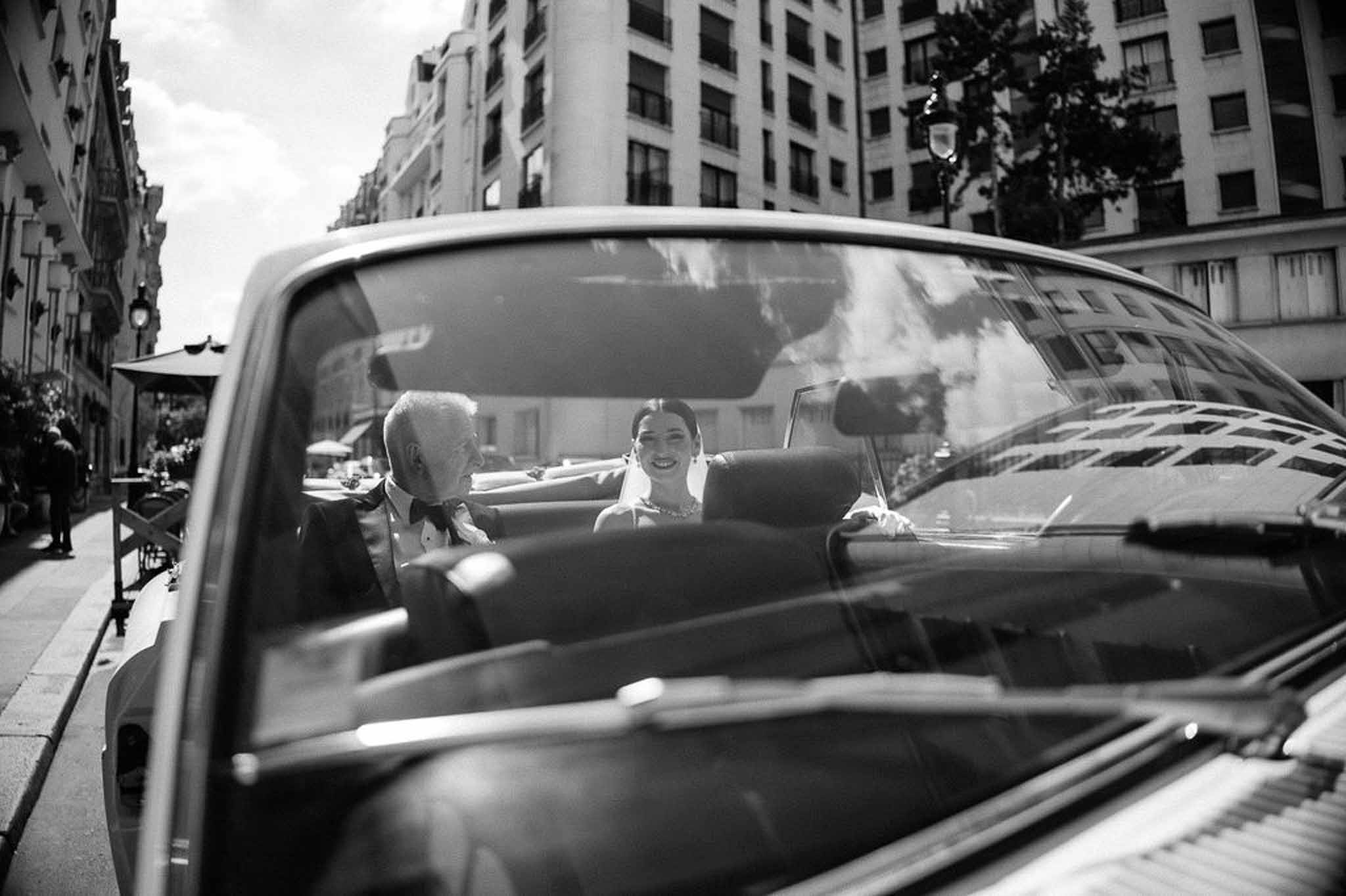 Bride and father laughing inside vintage convertible on Haussmann street through windshield in B&W