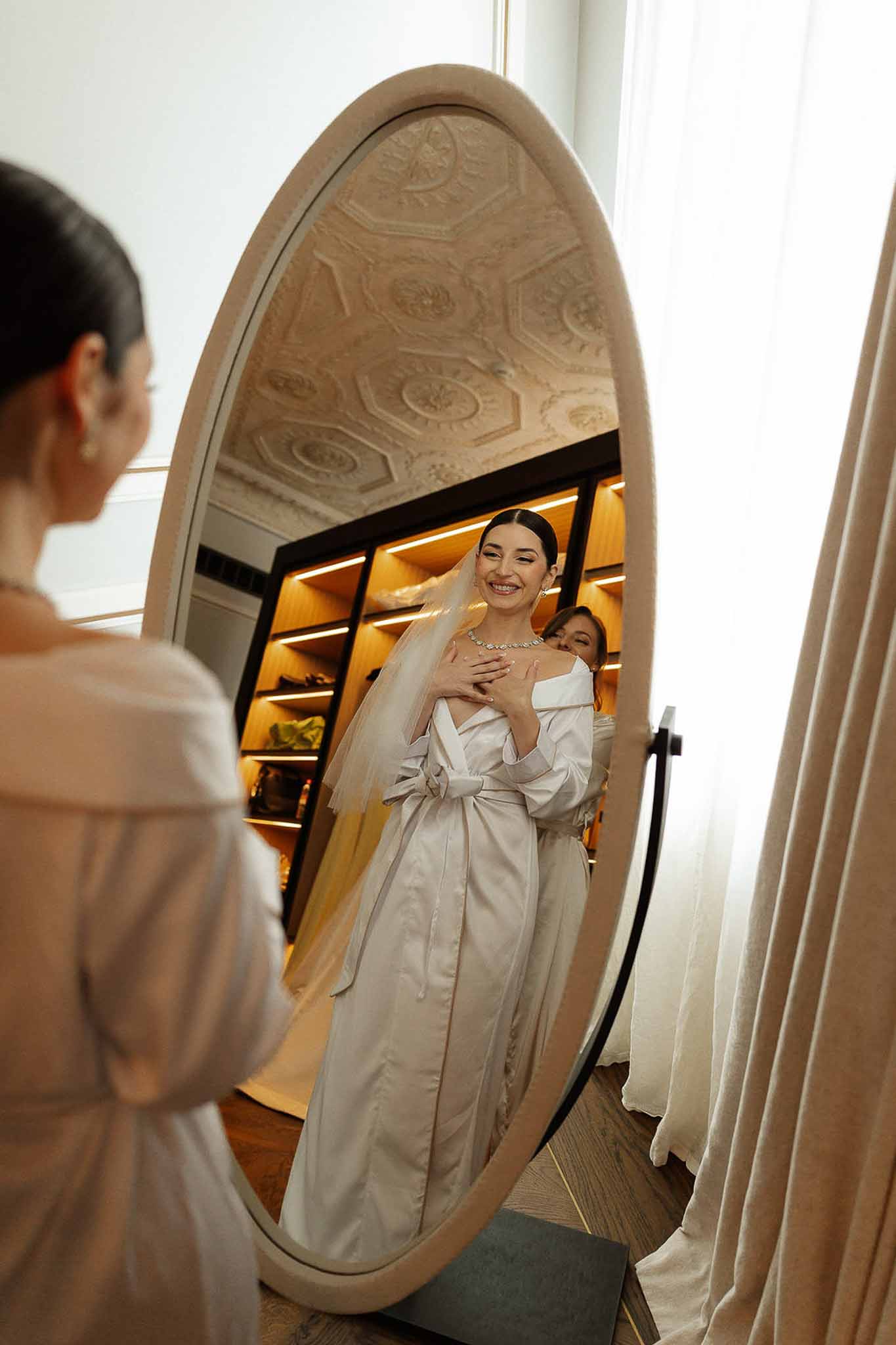 Bride in off-shoulder satin gown with cathedral veil and diamond necklace reflected in oval freestanding mirror