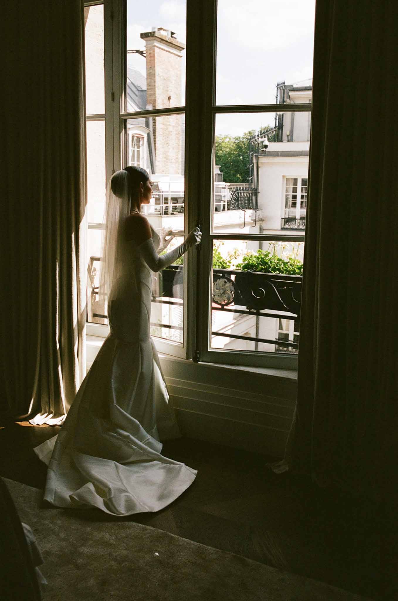 Bride in off-shoulder mermaid gown silhouetted at Parisian window with Haussmann rooftops beyond