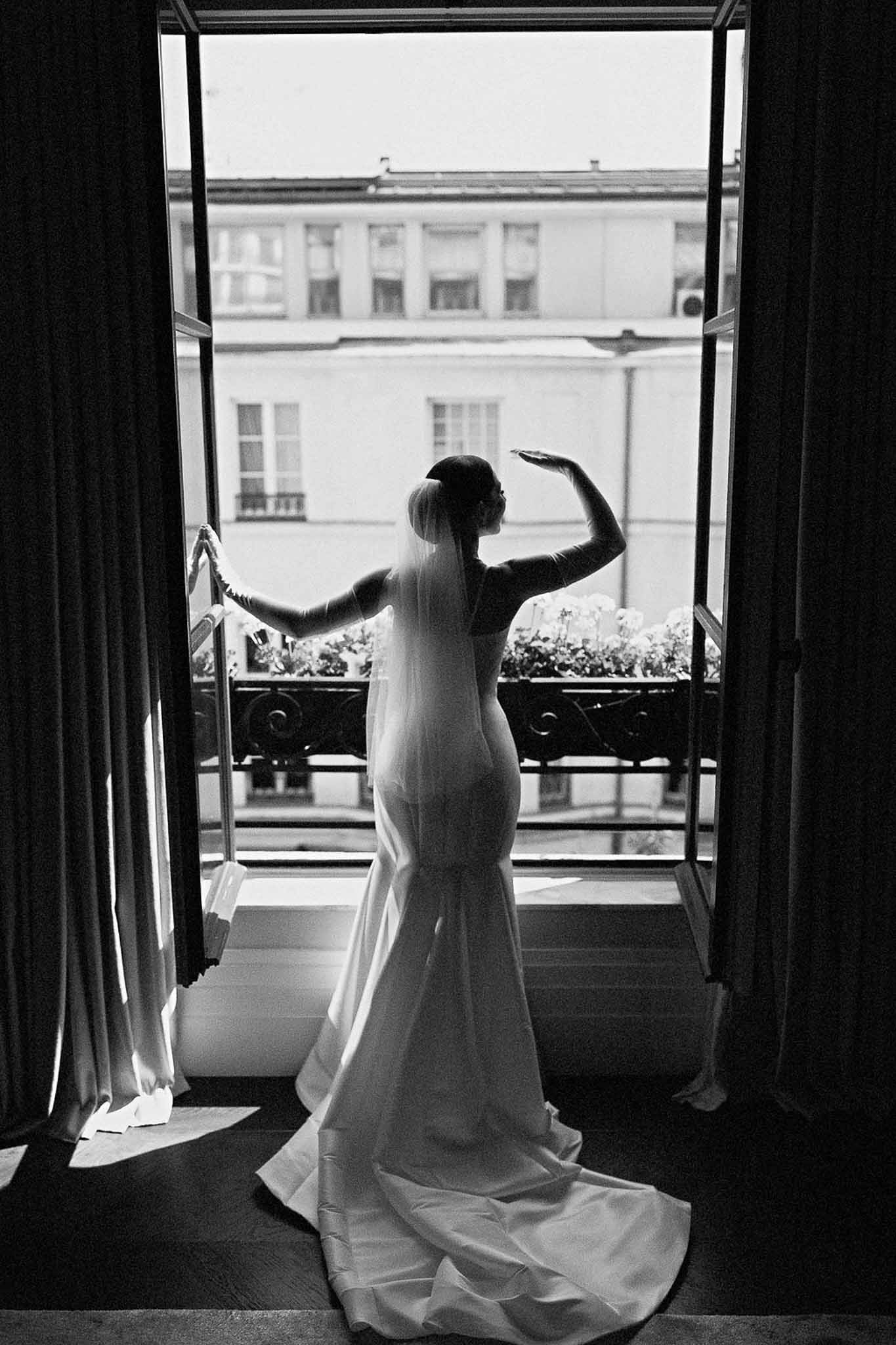 Bride silhouetted in mermaid gown at Parisian balcony French doors with Haussmann building across street in B&W