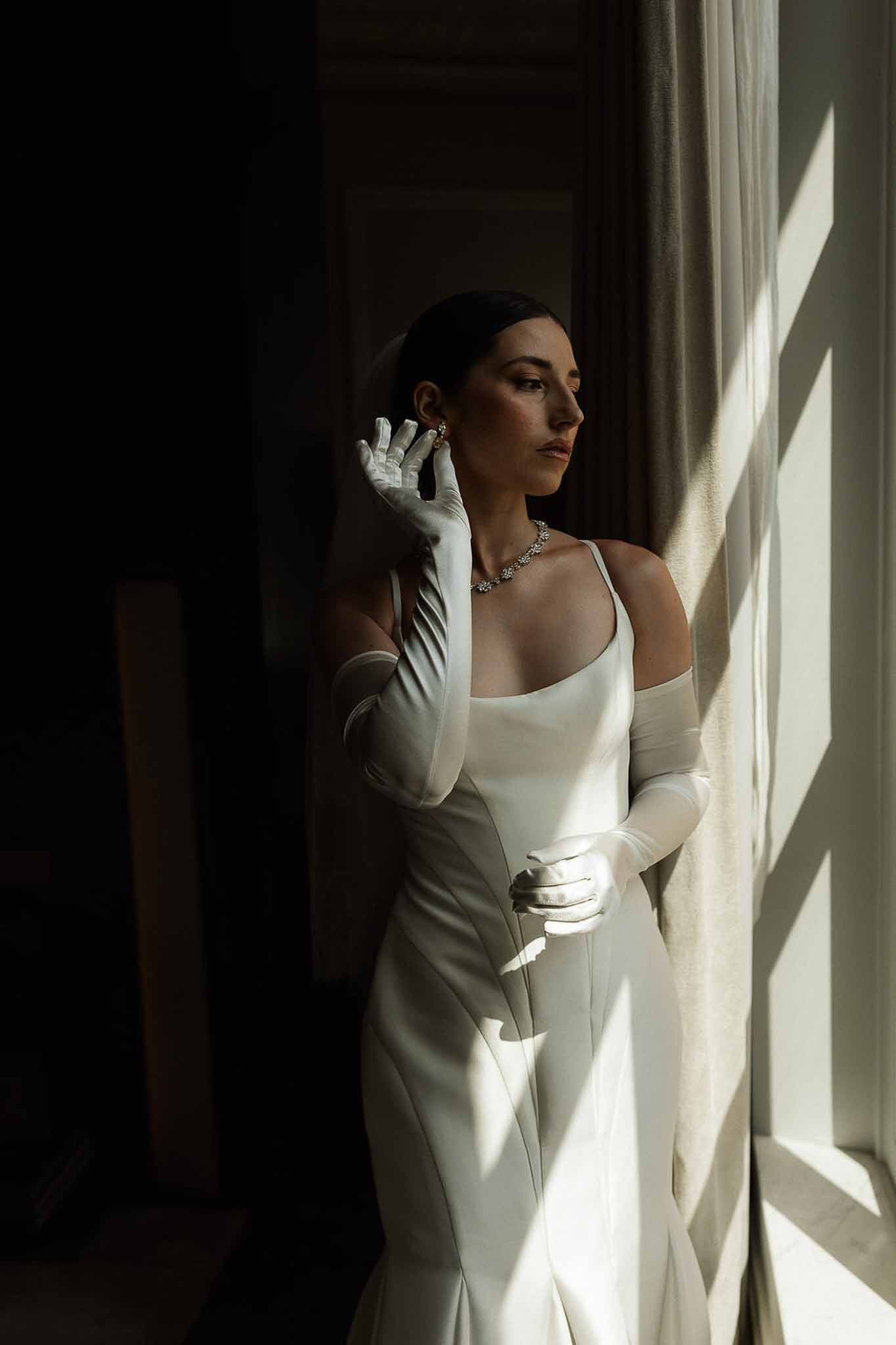 Bride in satin gown and elbow-length gloves with crystal necklace lit by dramatic window light