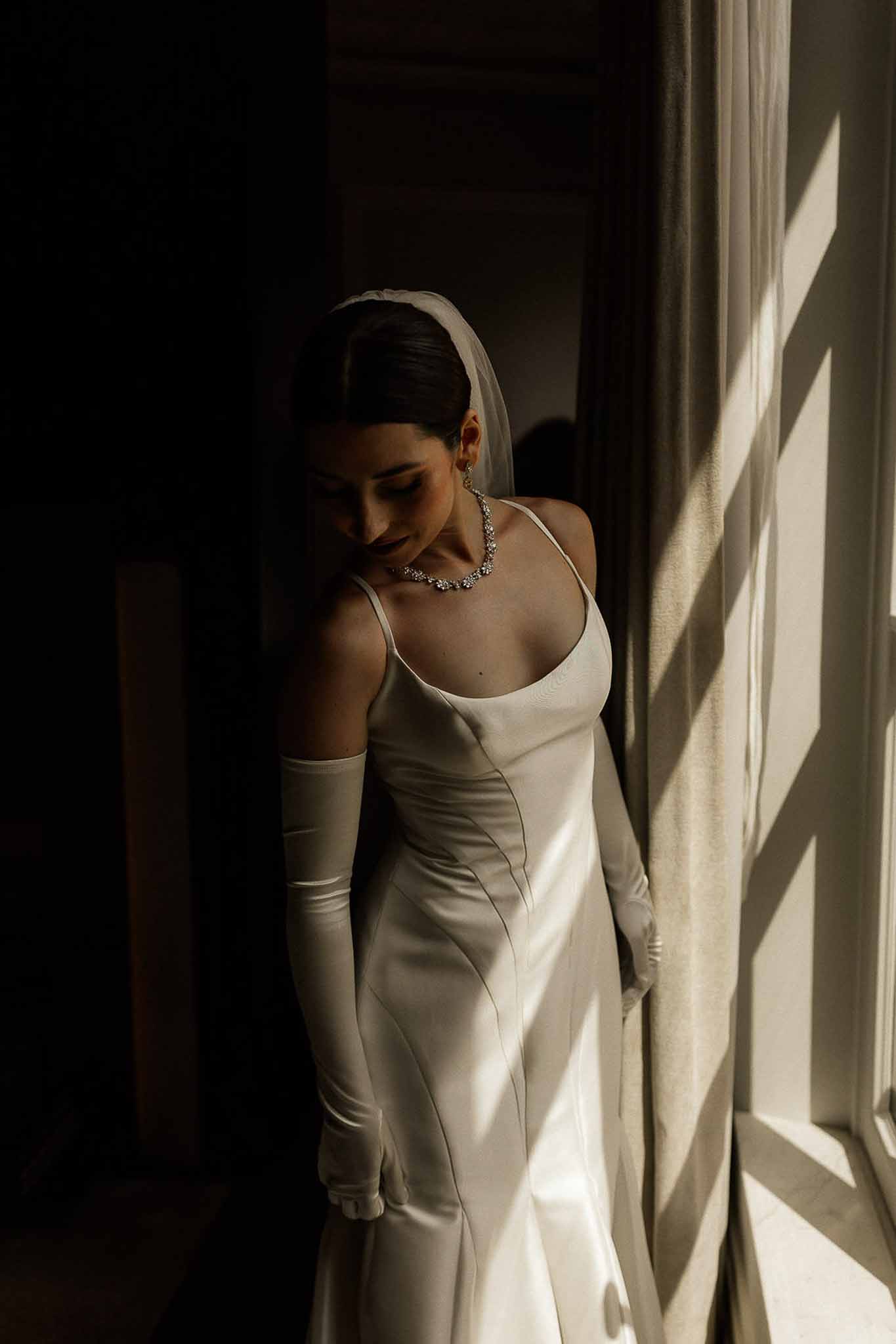 Bride in ivory satin slip gown with opera gloves and cathedral veil standing by window with geometric shadow patterns