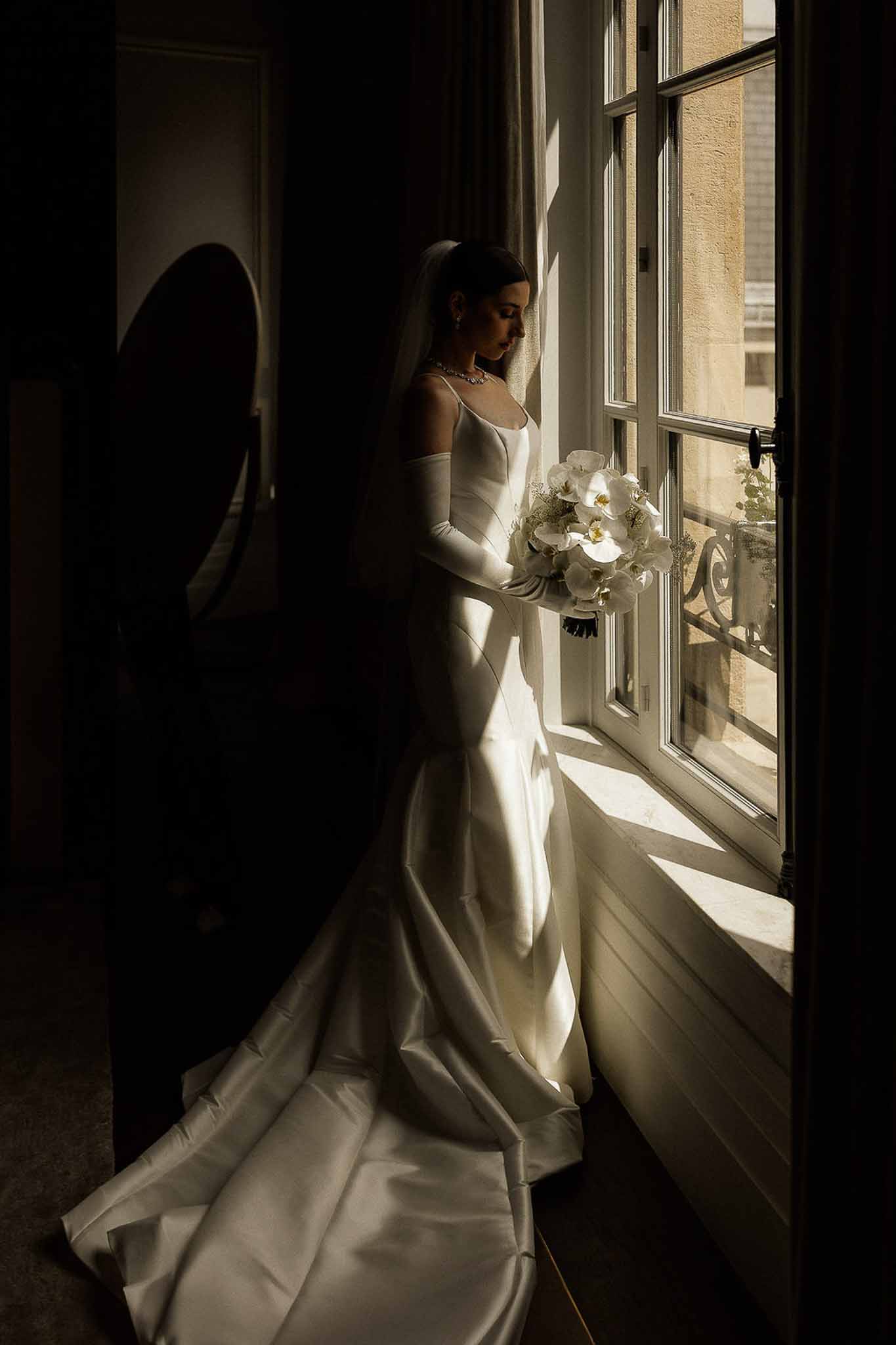 Bride in white satin mermaid gown with cathedral veil, long gloves, and orchid bouquet beside a tall French window