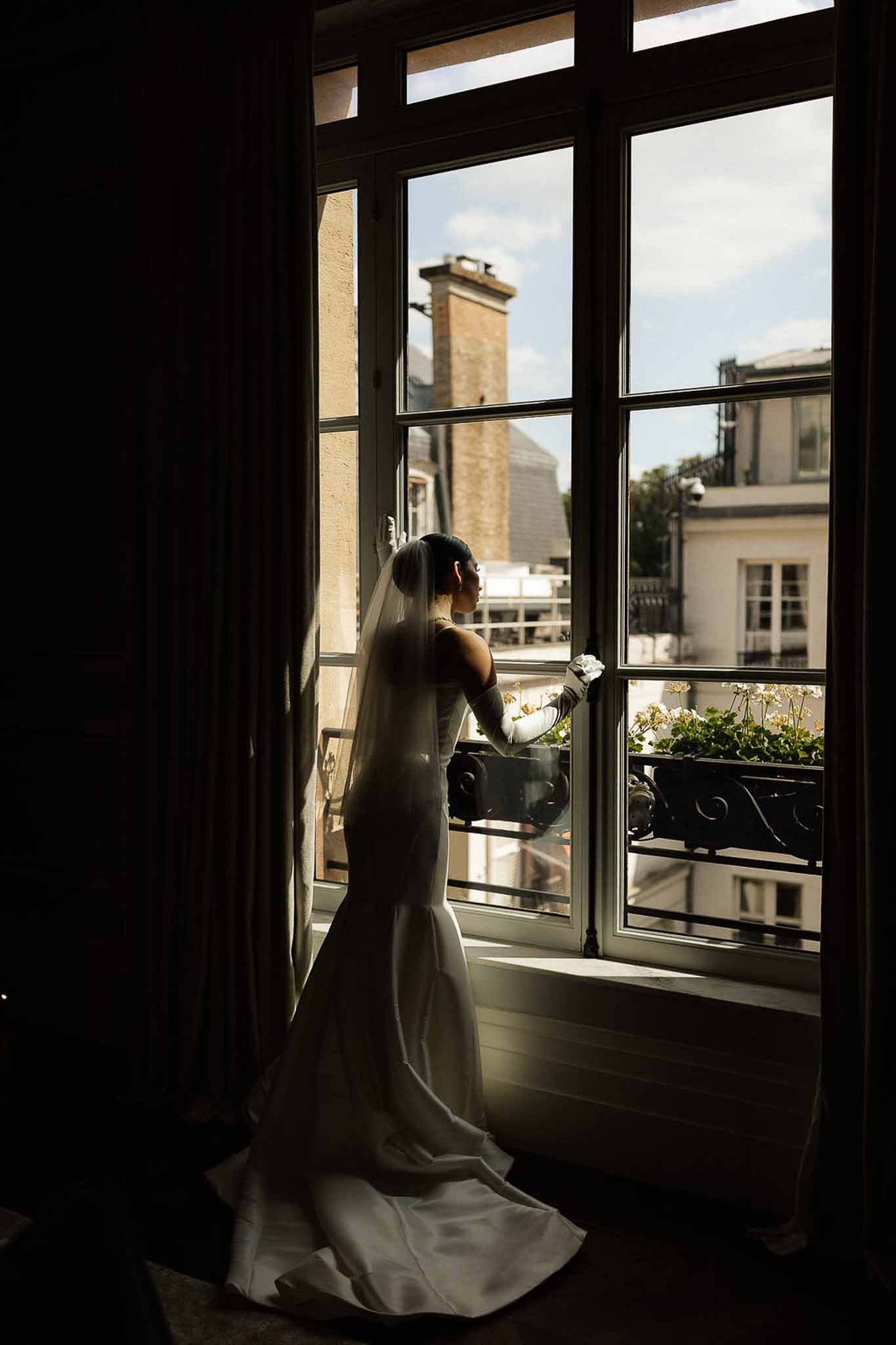 Bride silhouetted at tall window in ivory satin mermaid gown with opera gloves overlooking Parisian rooftops