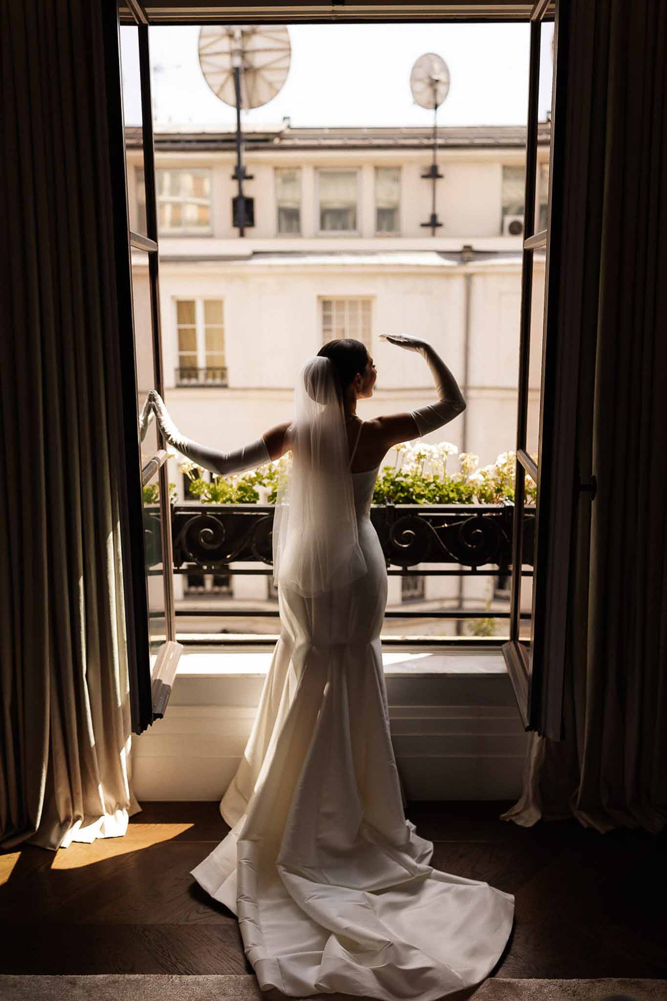 Bride from behind in ivory mermaid gown with cathedral train and gloves at open Parisian balcony doors