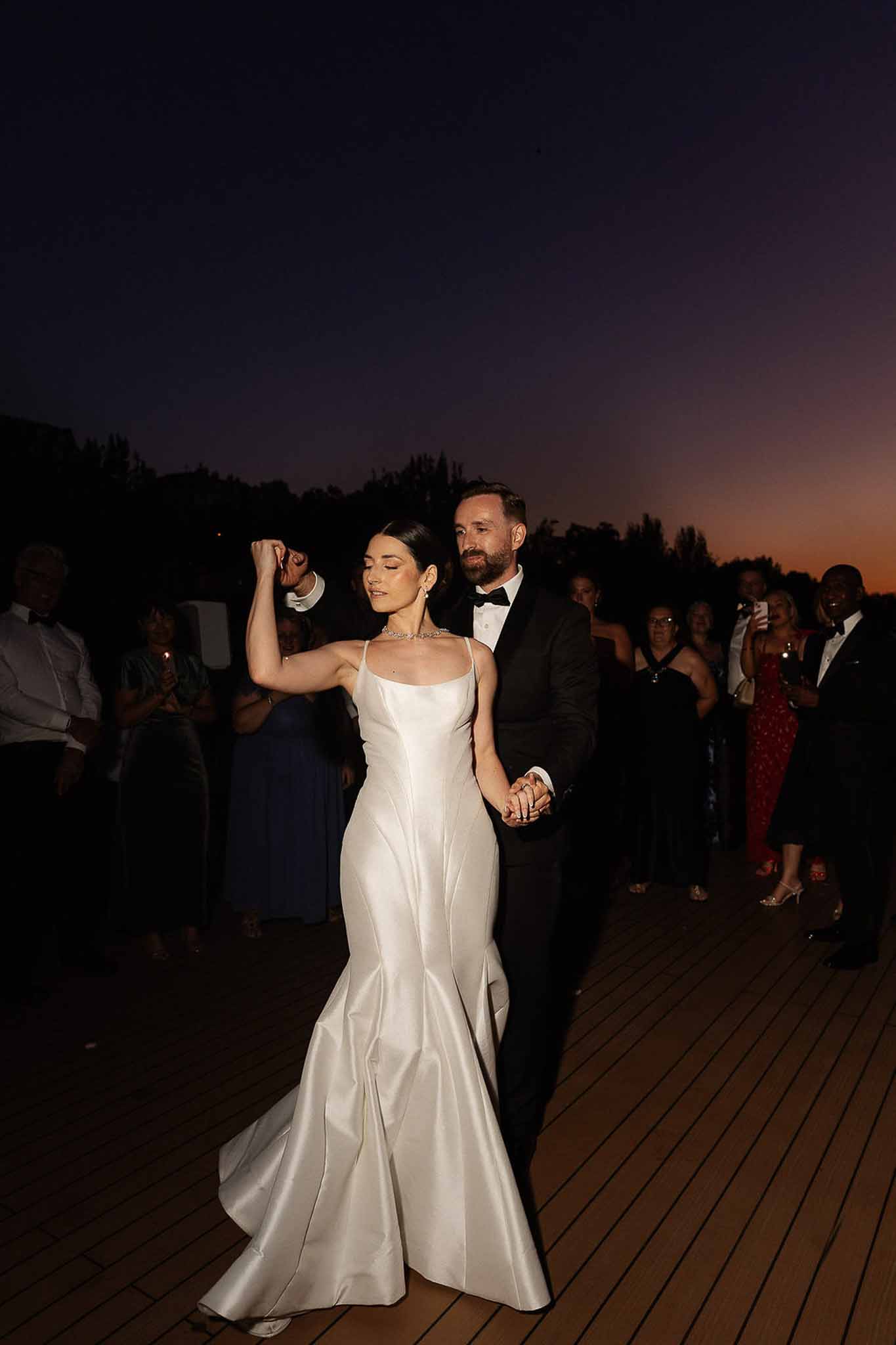 Bride and groom first dance on outdoor deck at dusk with sunset sky and watching guests
