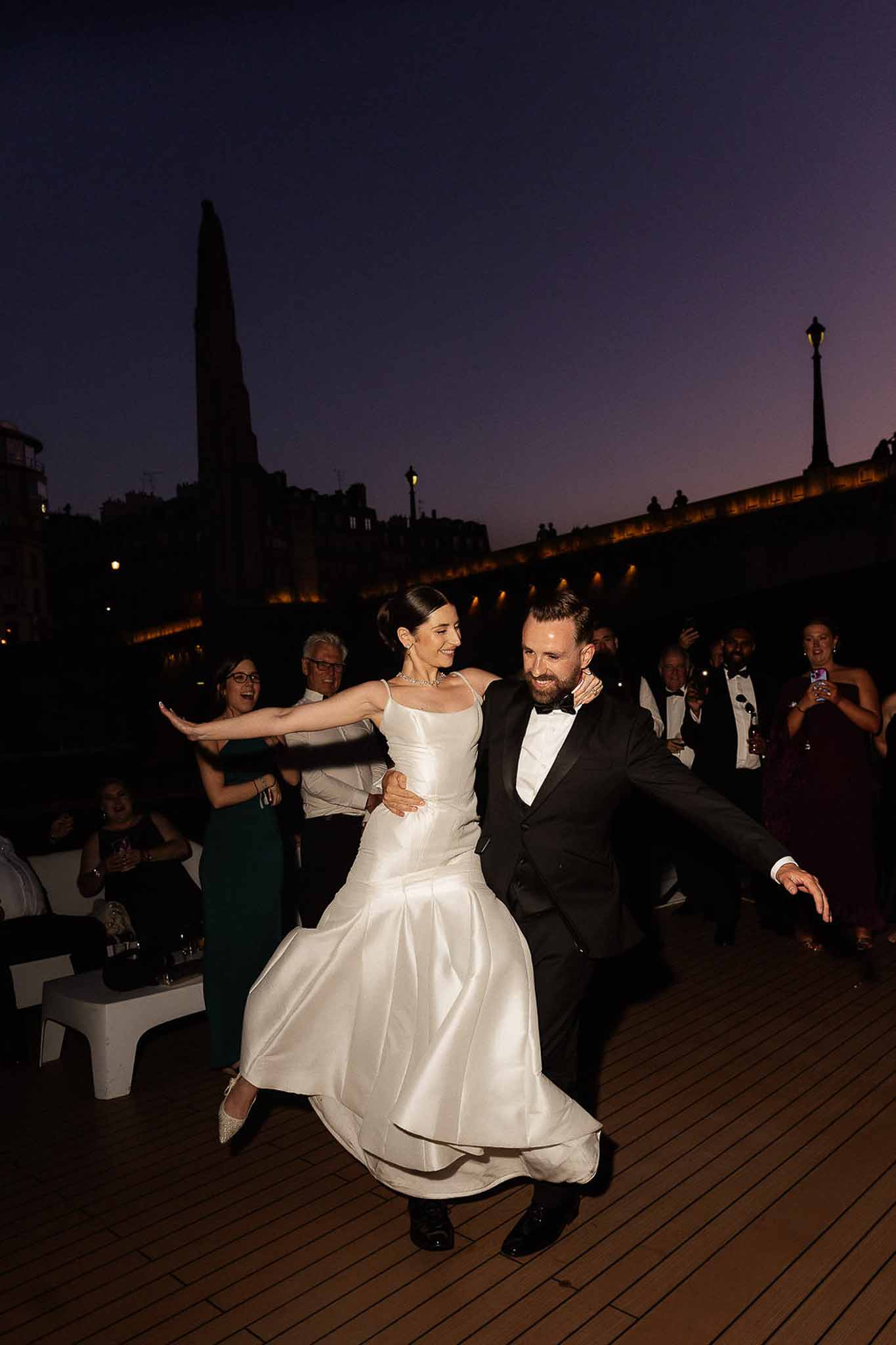 Couple first dance on Paris river boat deck at dusk with illuminated bridge and guests cheering behind