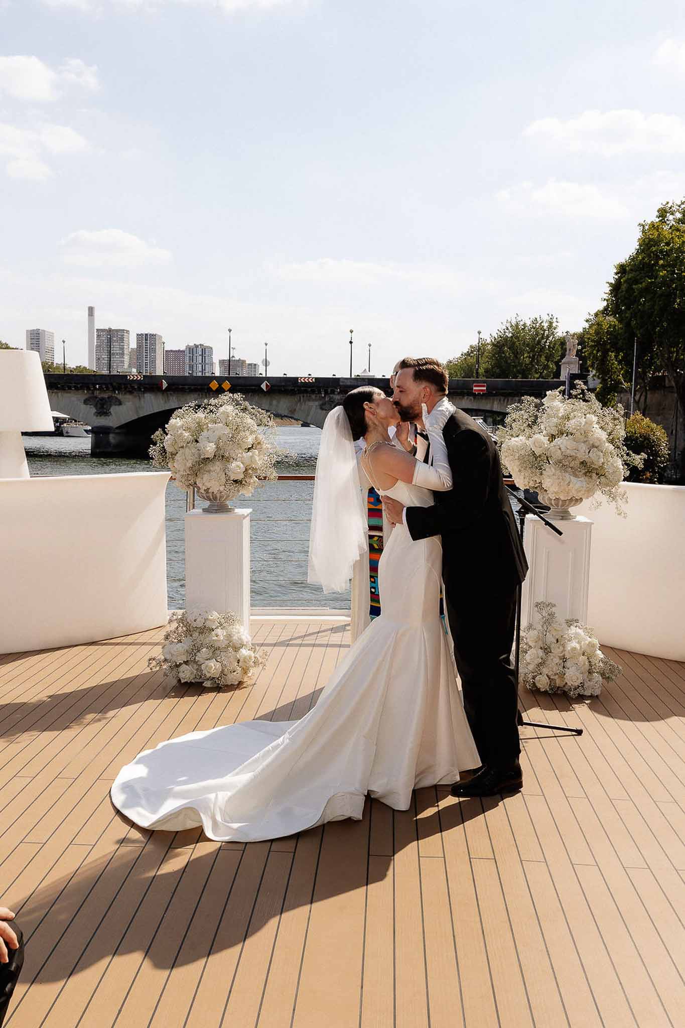 First kiss on Seine riverboat deck with white rose pedestal columns and Paris bridge in background