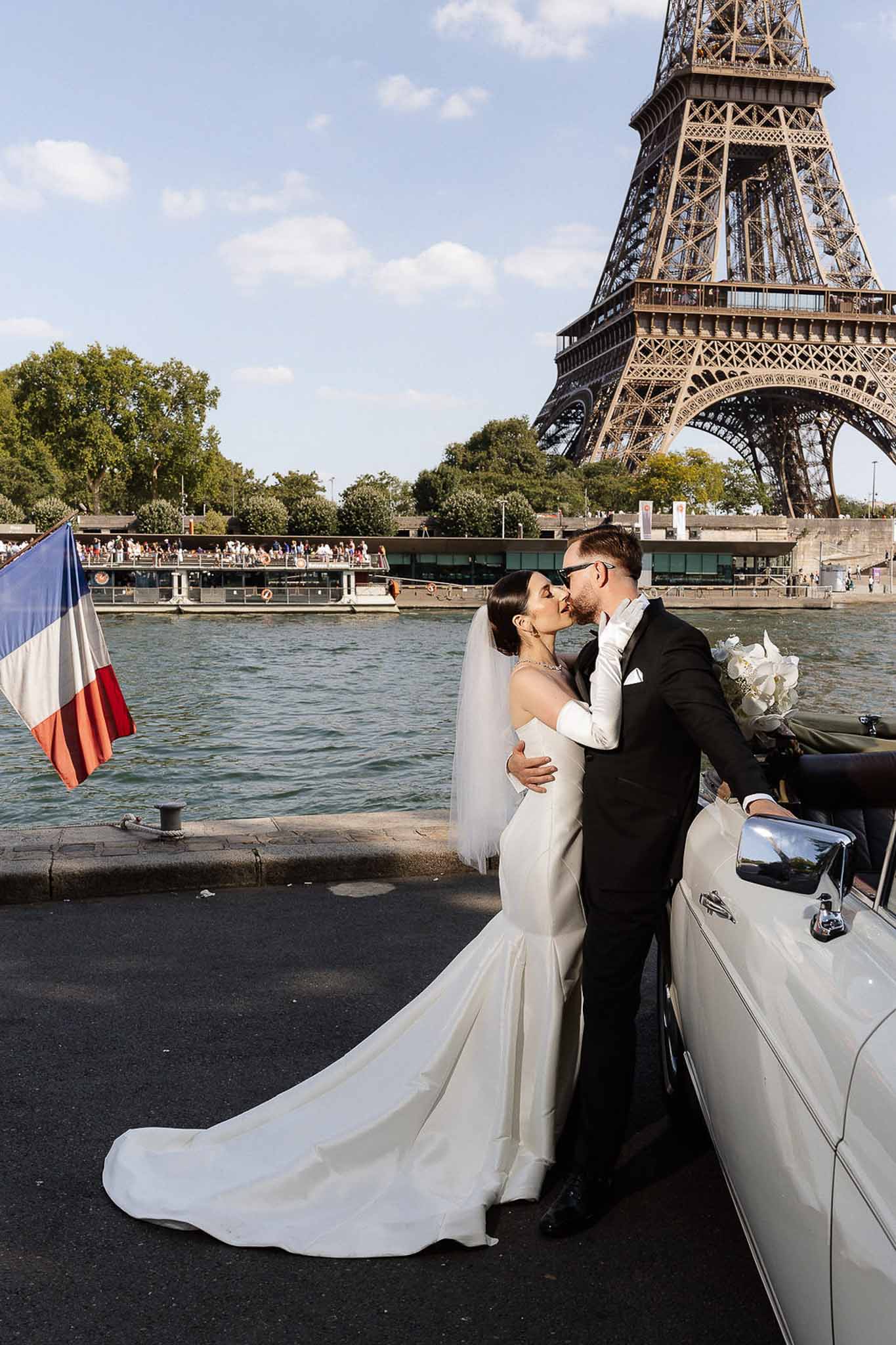 Couple kissing by Seine with Eiffel Tower, bride in ivory mermaid gown with gloves and vintage convertible beside