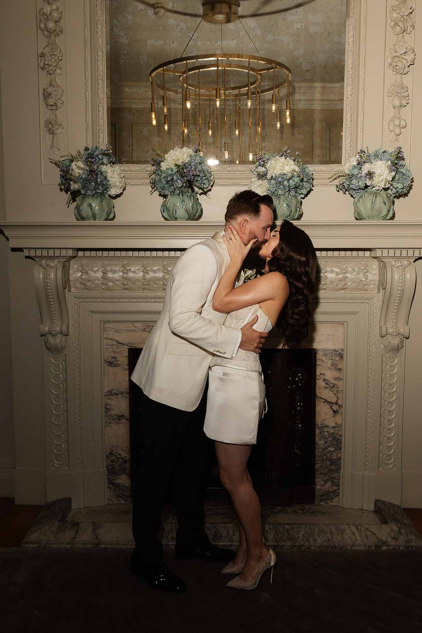 Bride and groom kissing in front of a marble fireplace decorated with white and blue hydrangea arrangements