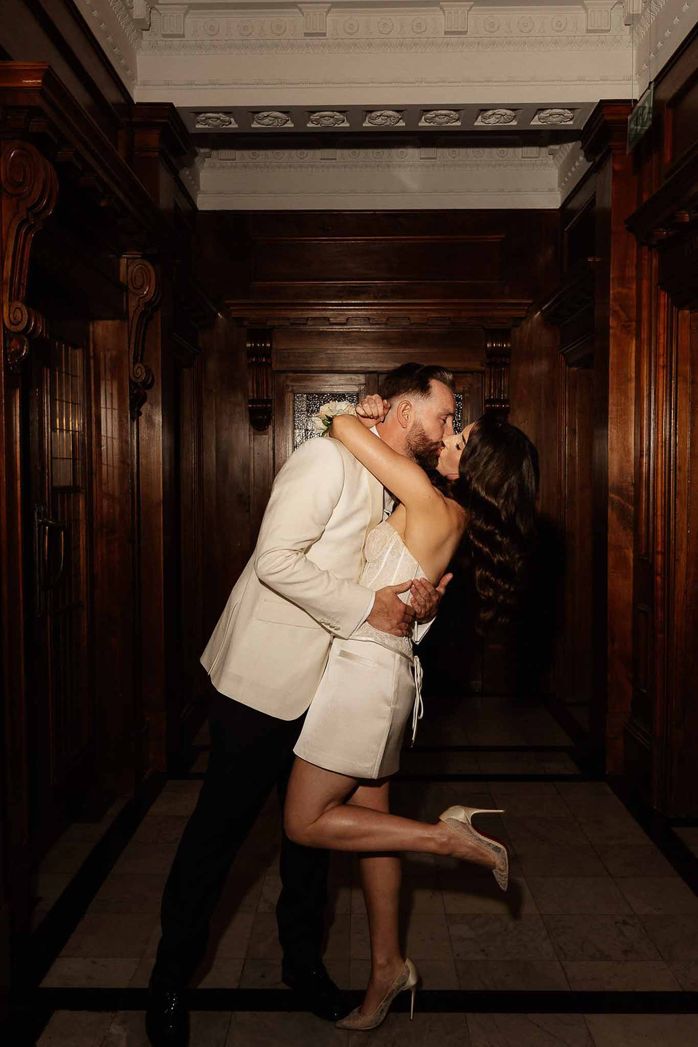 Dip kiss in mahogany-panelled corridor with bride in lace mini dress and red-sole stilettos