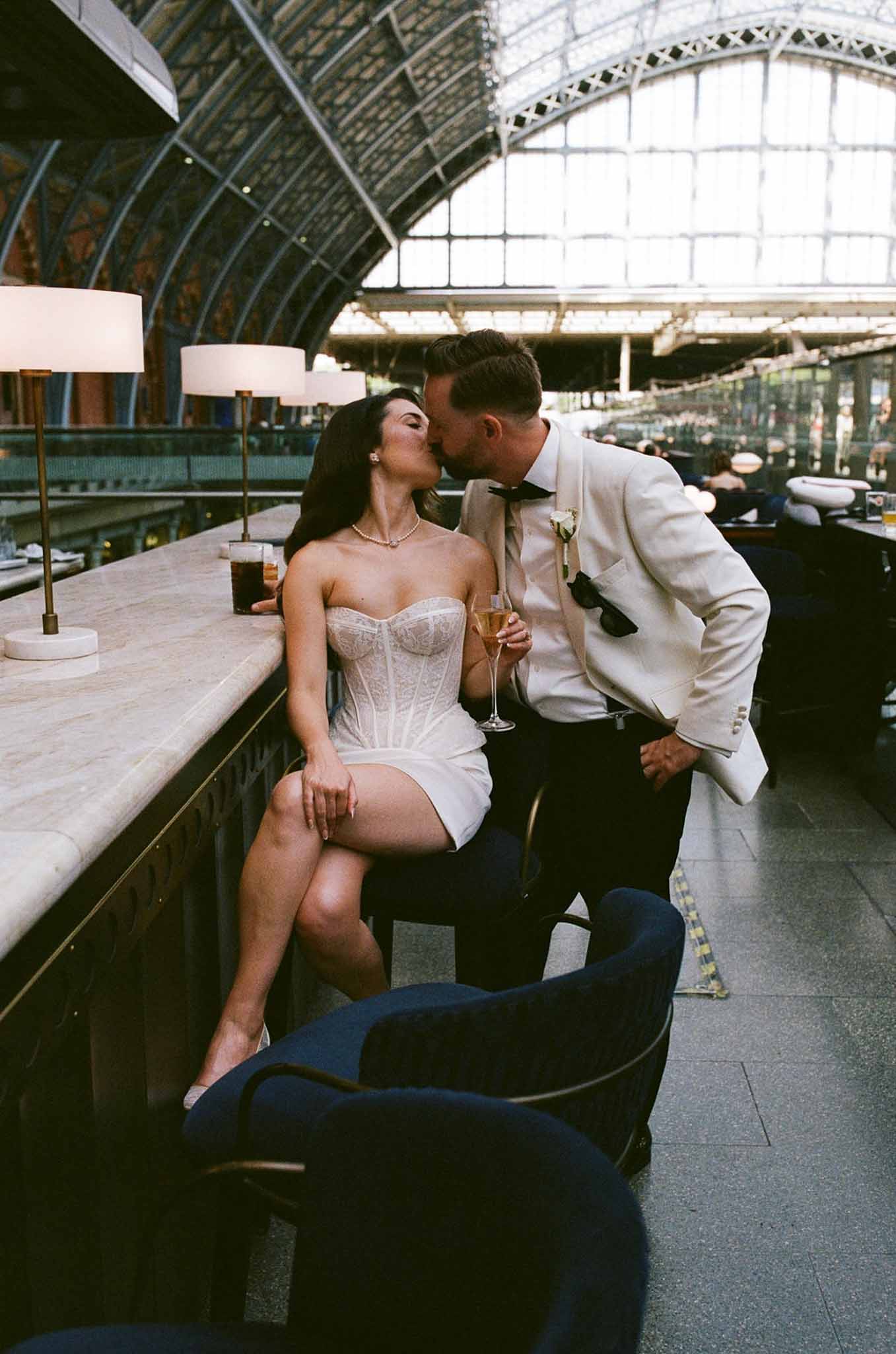 Bride in ivory lace mini dress and groom in cream dinner jacket kissing at marble bar in grand train station