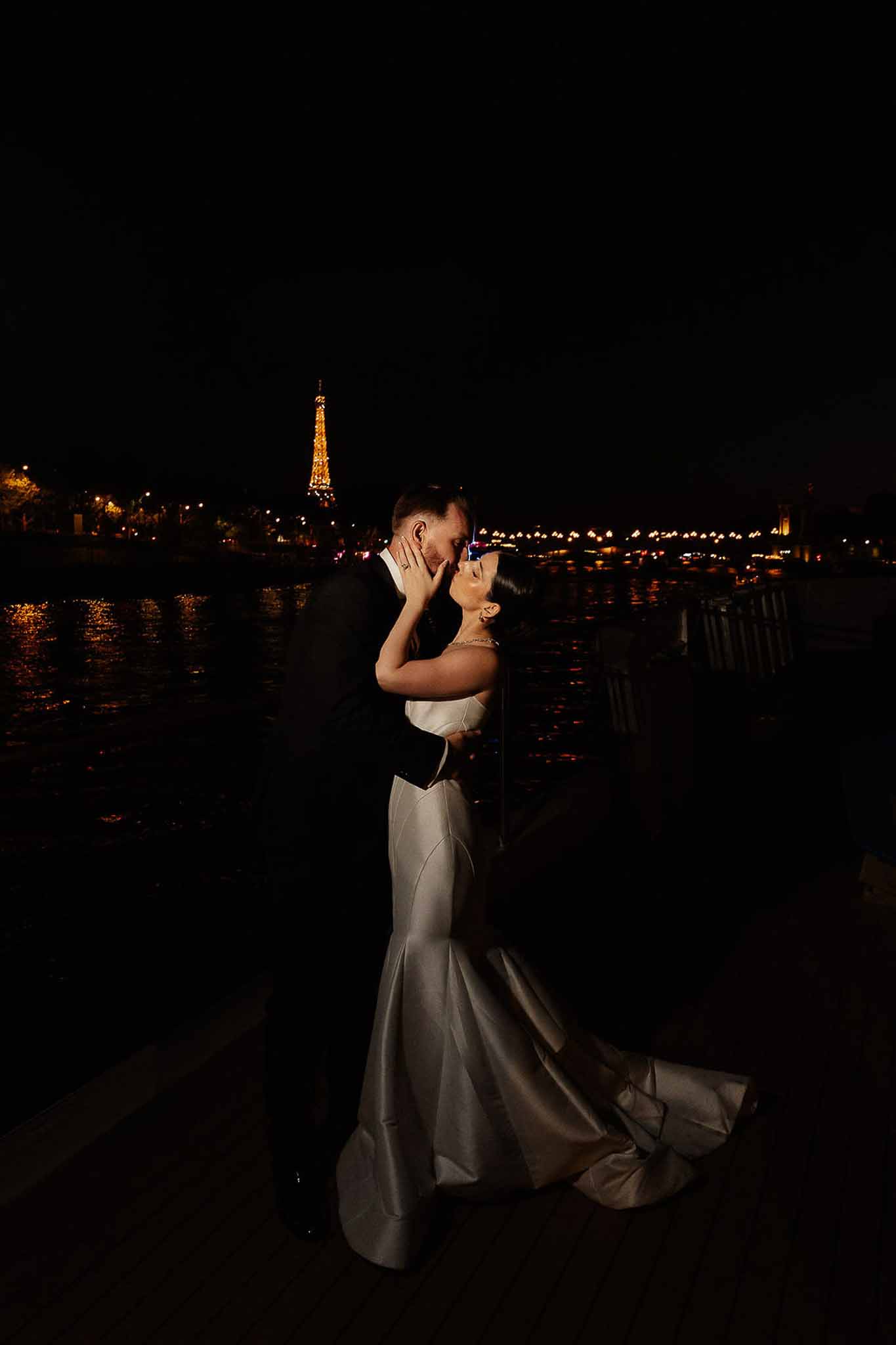 Bride and groom kissing at night along Seine riverbank with illuminated Eiffel Tower in background
