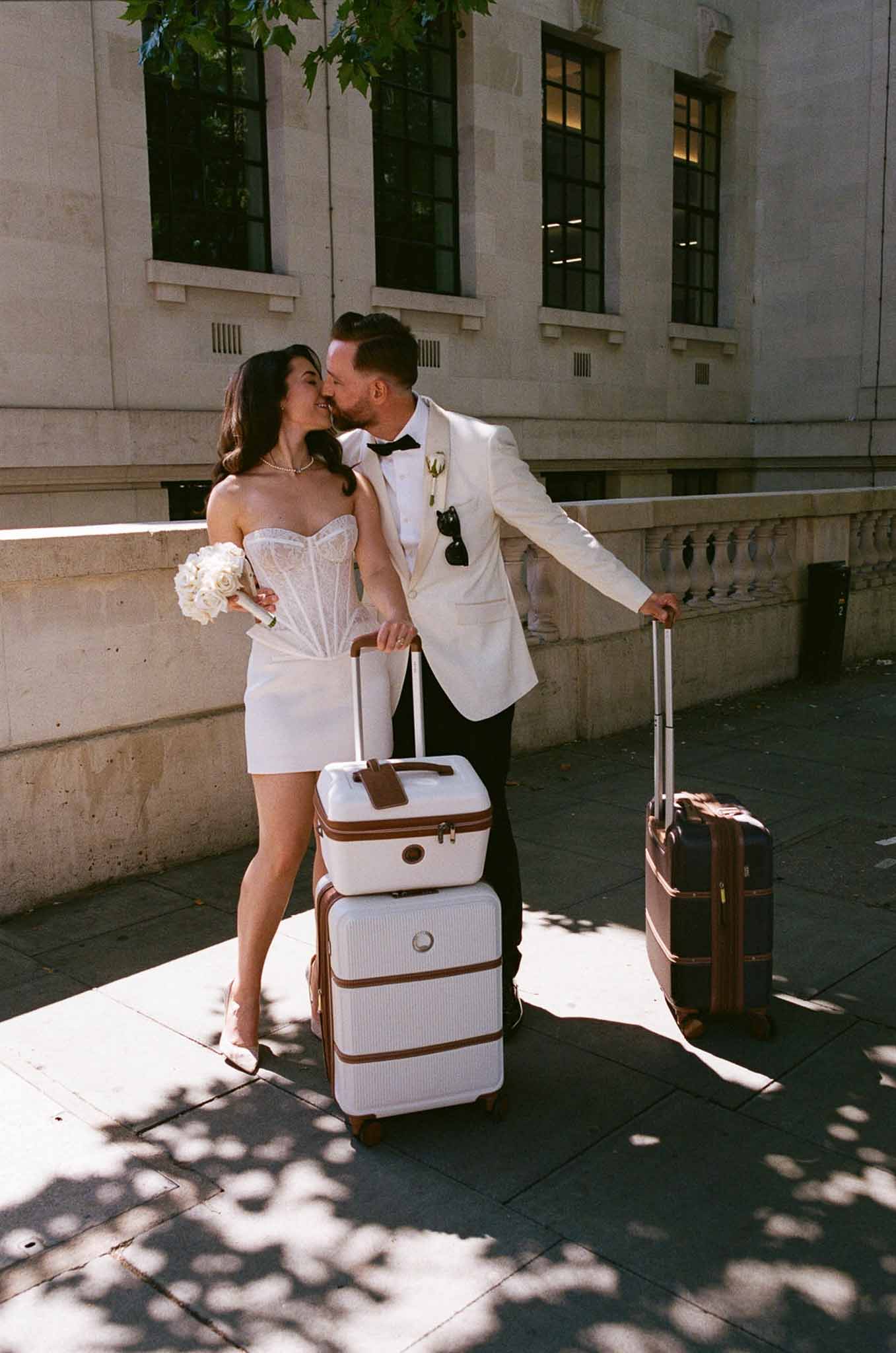 Bride and groom kissing on city pavement with vintage luggage, bride in white mini skirt and lace top
