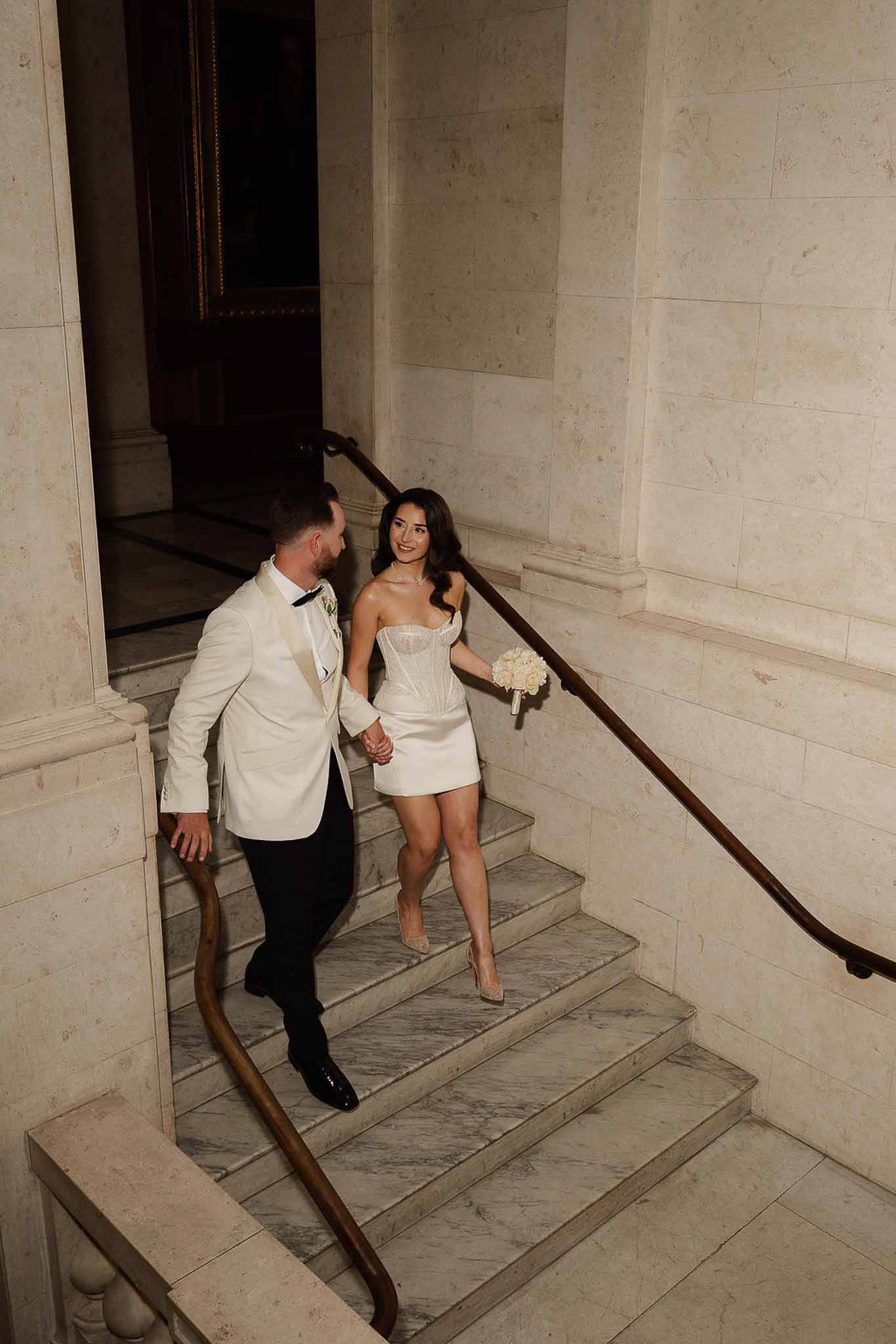 Overhead shot of couple descending marble staircase, bride in white corset mini dress with ivory rose bouquet