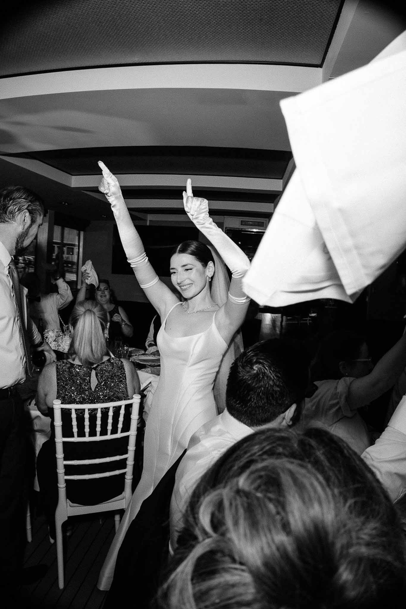 Black-and-white photo of bride dancing with arms raised while guests wave napkins at indoor evening reception