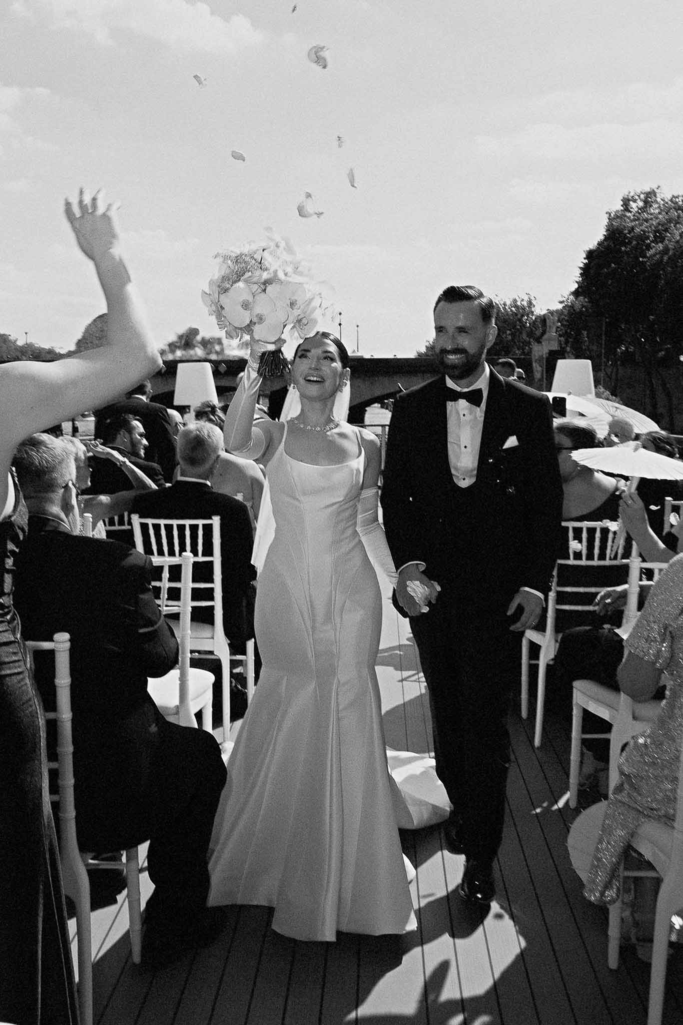Black and white couple walking up aisle with orchid bouquet raised and petals thrown by guests with white parasols
