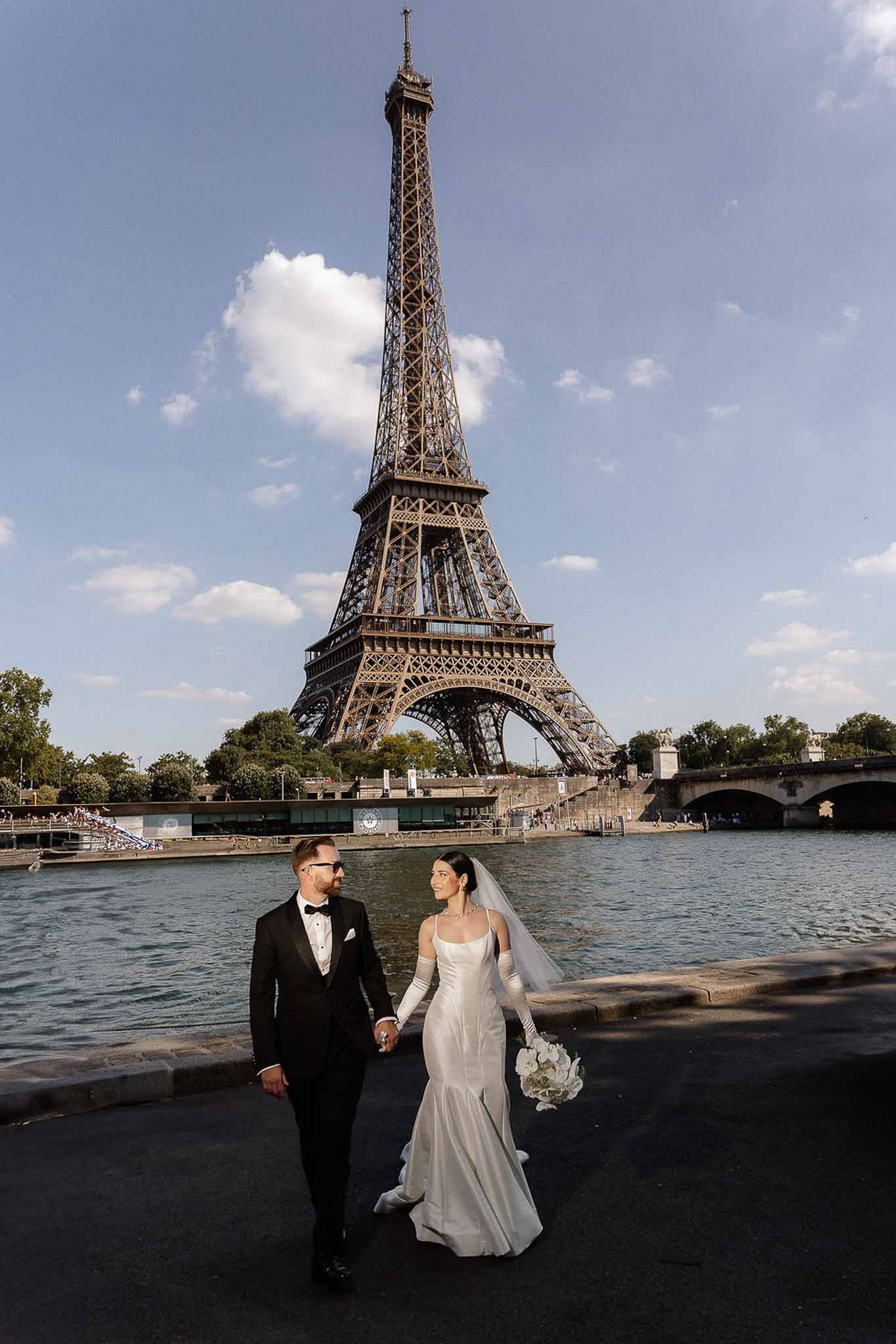 Bride and groom walking hand in hand along the Seine with the Eiffel Tower behind them in Paris