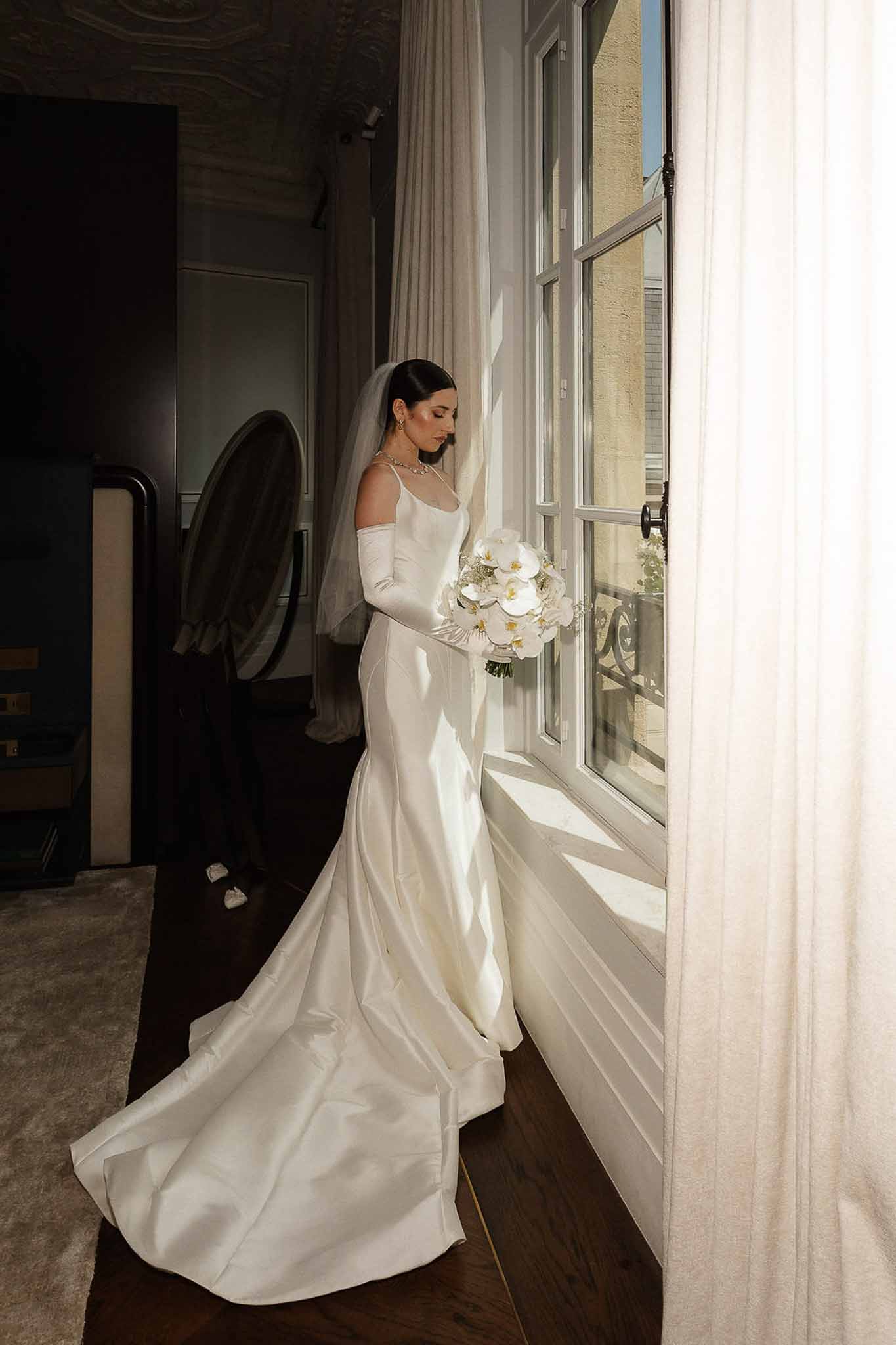 Bride in ivory satin mermaid gown with cathedral veil and opera gloves holding white orchid bouquet by window
