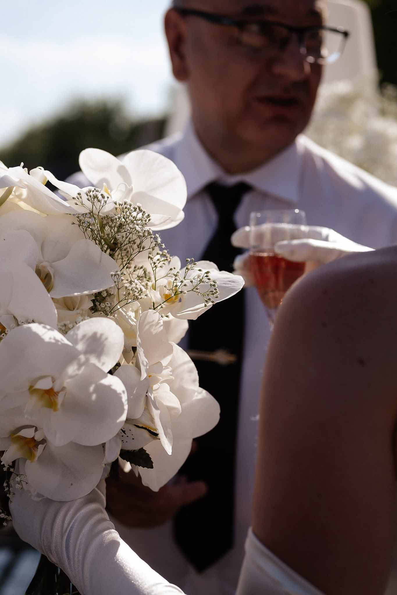 Close-up of bride holding white orchid and baby's breath bouquet with glass of rose champagne in gloved hand