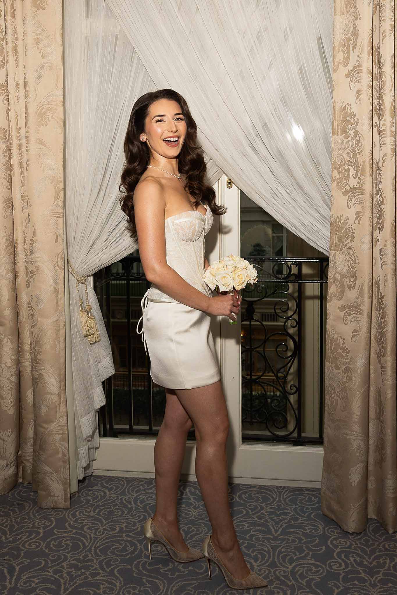 Bride in white strapless mini dress with pearl choker laughing by window with gold damask curtains