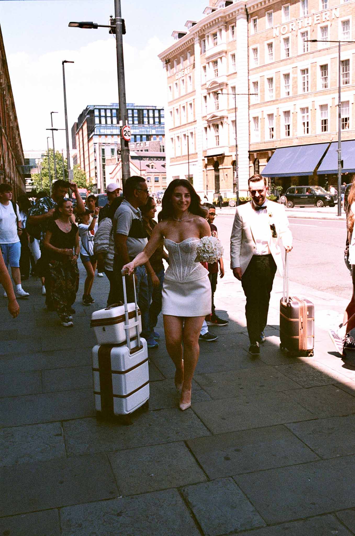 Bride in white corset mini and groom in white tuxedo pulling suitcases on urban pavement shot on film