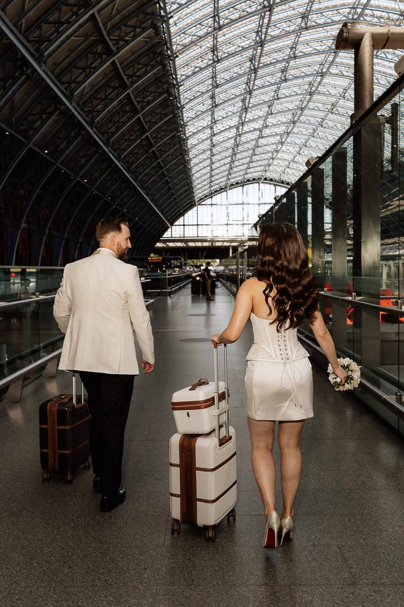 Bride and groom walking through train station with matching luggage, shot from behind