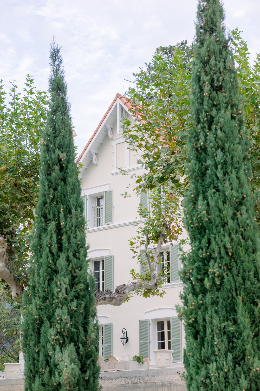 Three-story French country house with cream walls, terracotta roof, and green shutters flanked by cypress trees