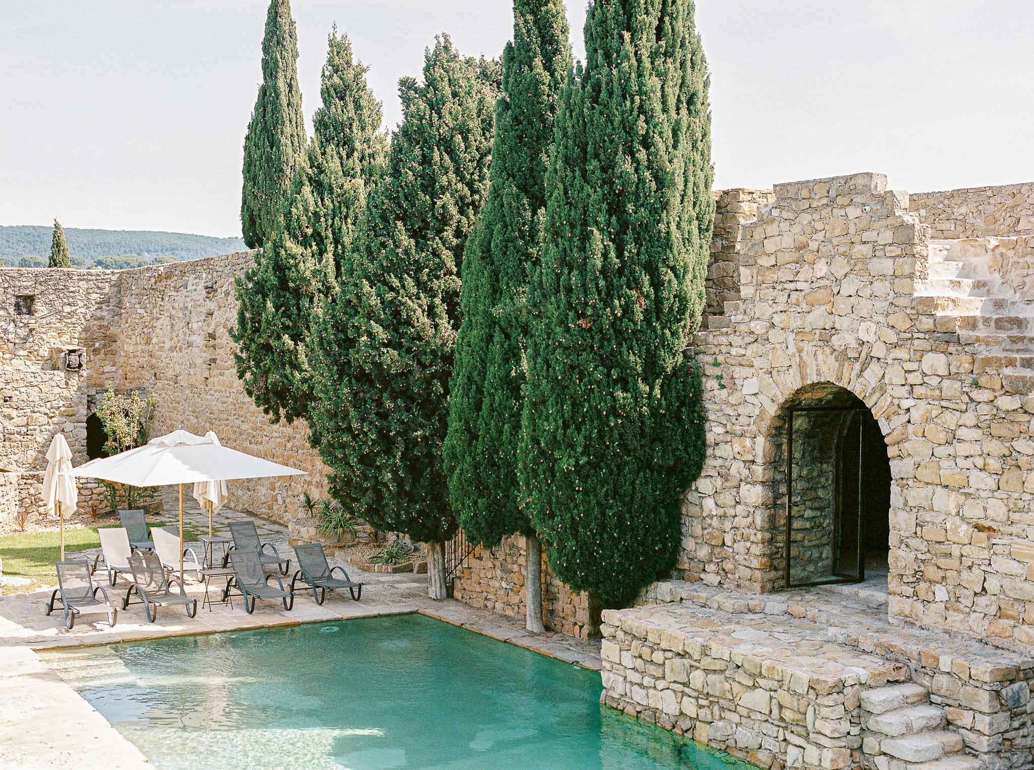 Limestone walled pool area with turquoise water grey loungers cream parasols cypress trees and stone archway with glass doors