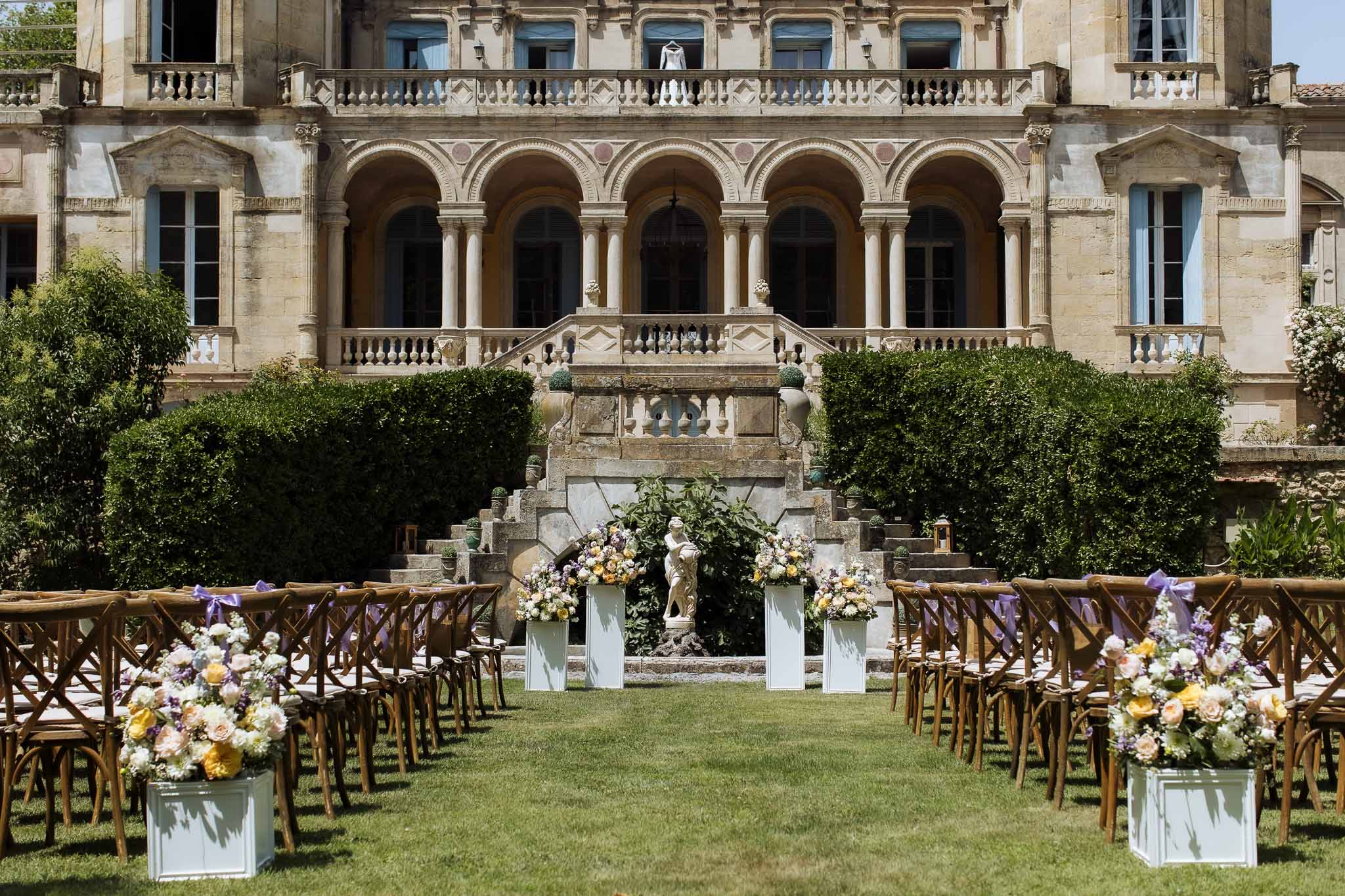 Empty ceremony setup with cross-back chairs and lavender ribbons facing chateau with floral pedestals