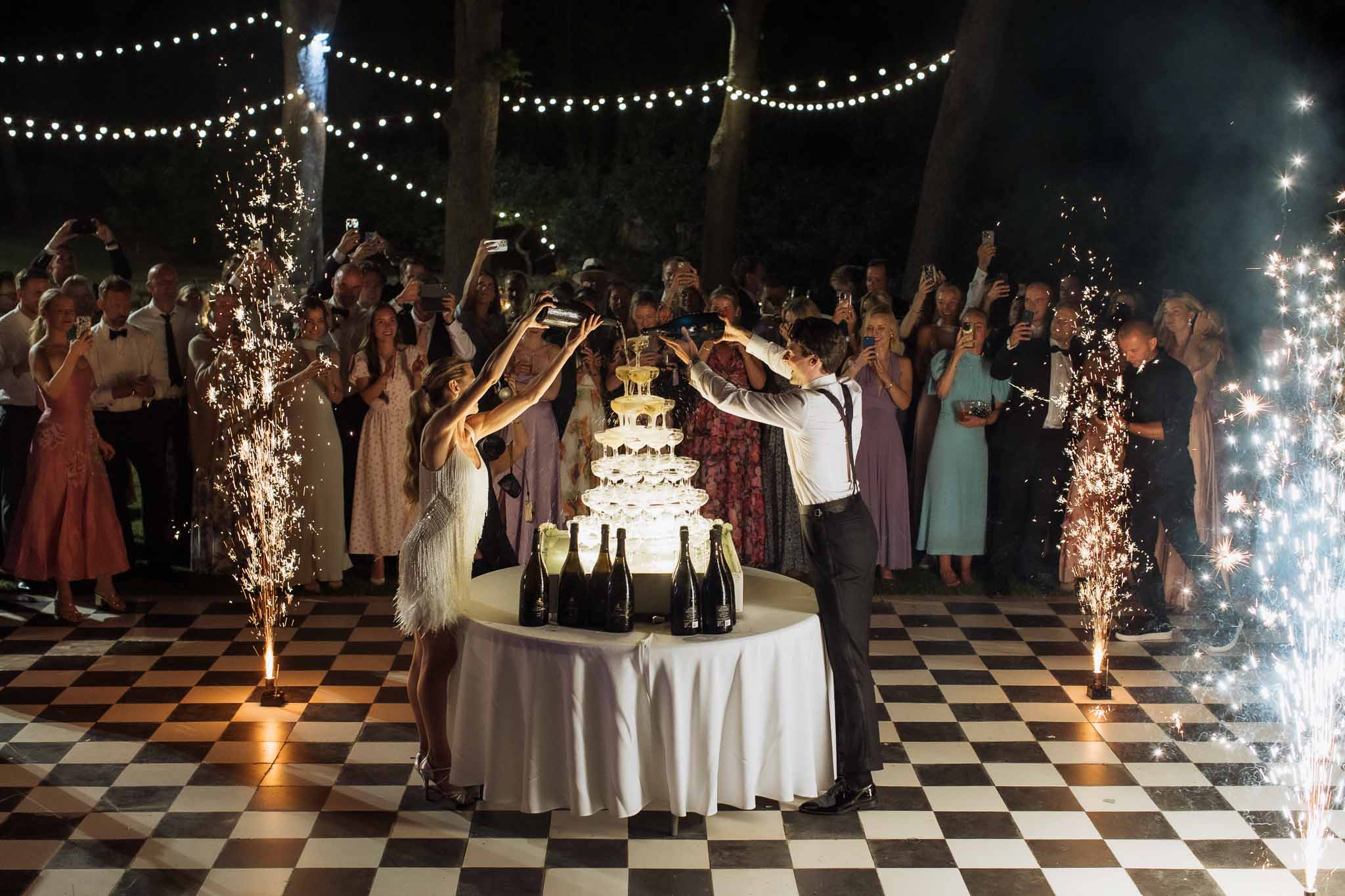 Couple pouring champagne tower on checkered dance floor with sparkler fountains and string lights at night