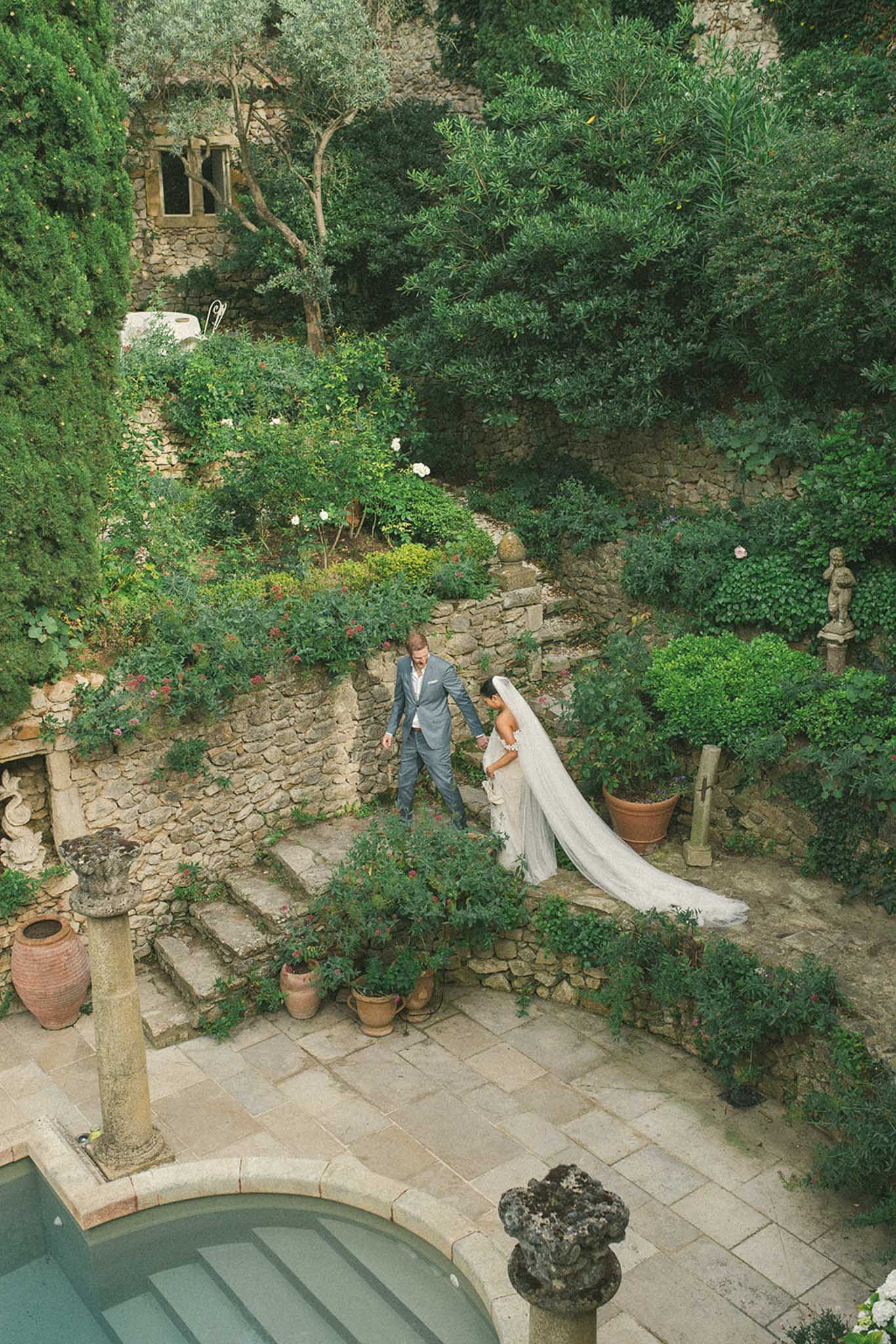 Aerial couple walking stone terrace steps with cathedral veil trailing beside turquoise pool at chateau