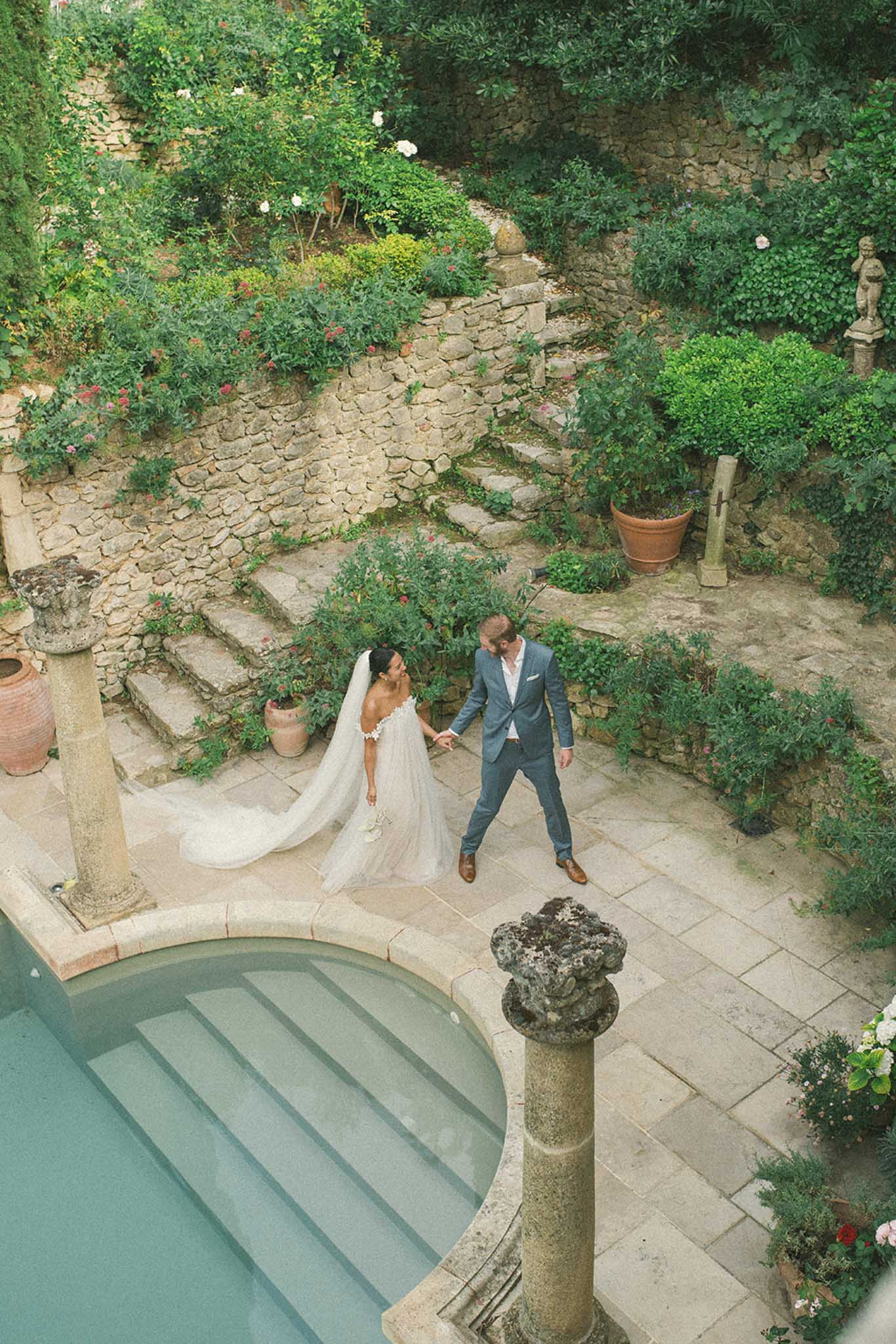 Overhead aerial view of bride and groom walking hand in hand on stone terrace beside turquoise pool in walled garden