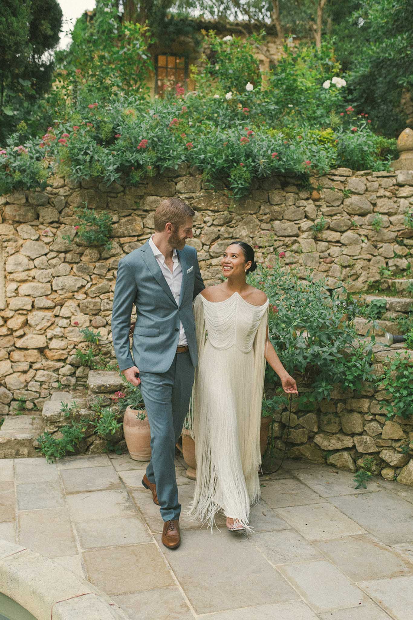 Couple in ivory fringe dress and slate blue suit walks smiling on stone terrace beside dry-stone wall