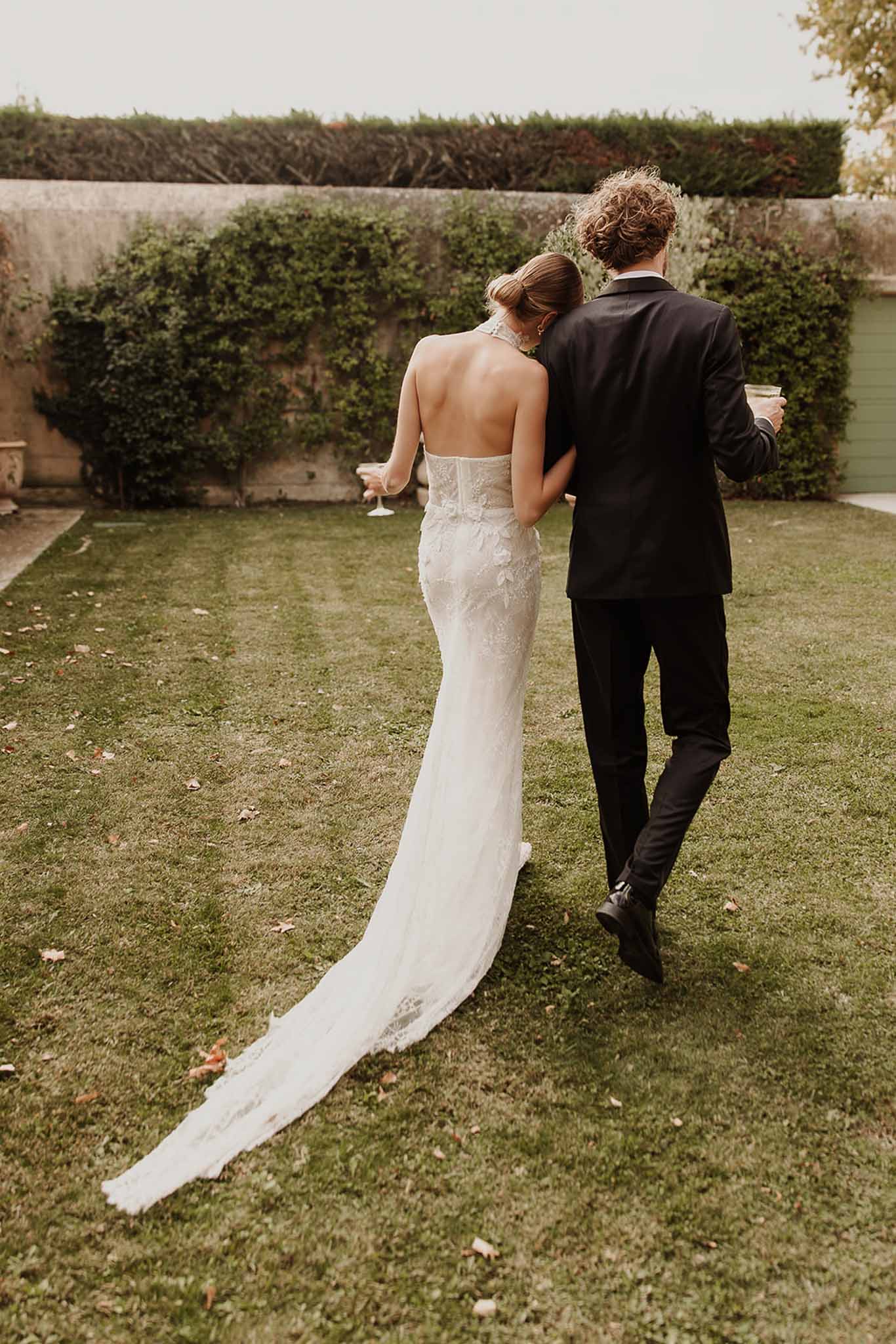Couple walking away across walled garden lawn holding champagne coupes in ivory lace gown