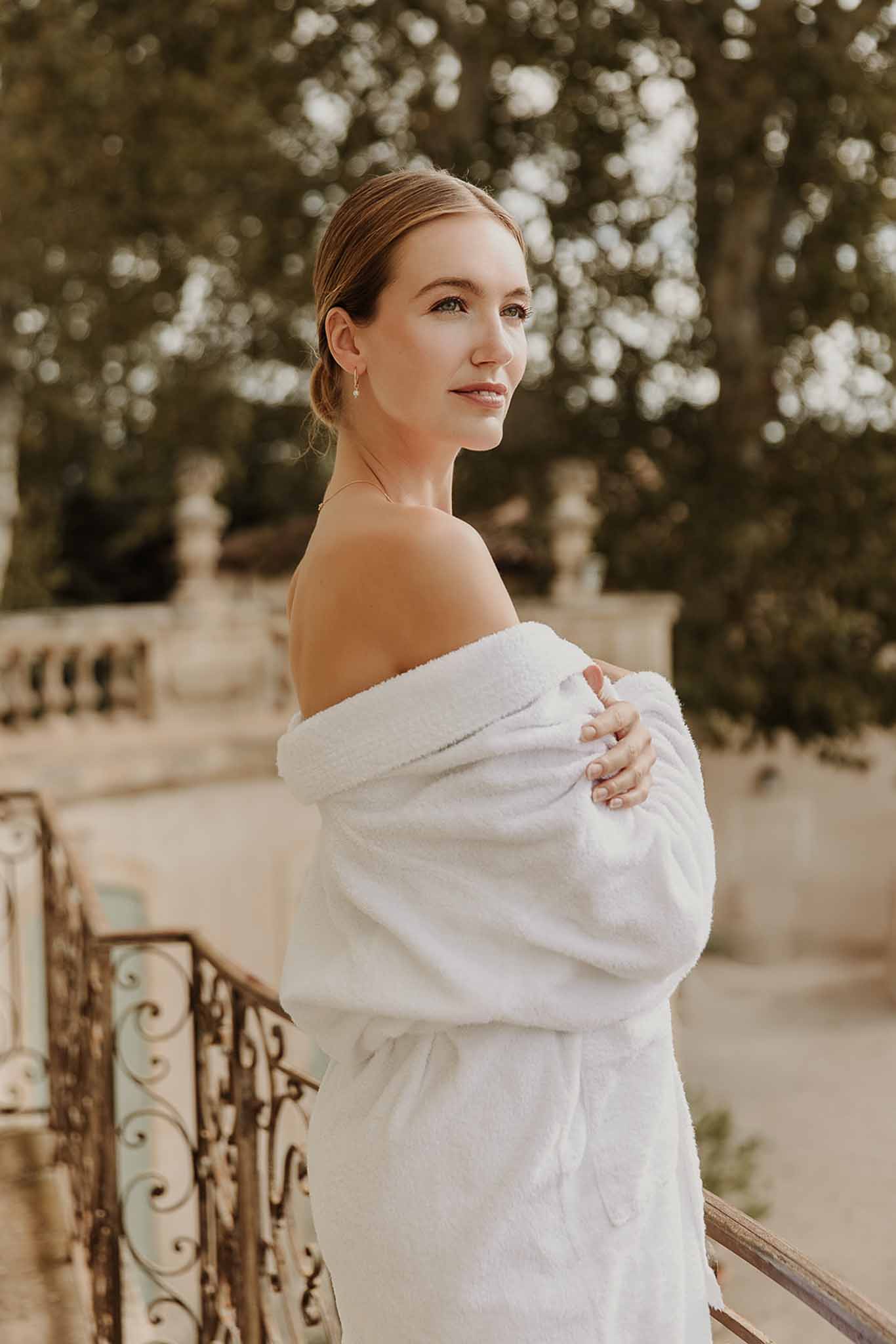 Bride in white robe on chateau terrace with sleek low bun and gold drop earrings looking over shoulder