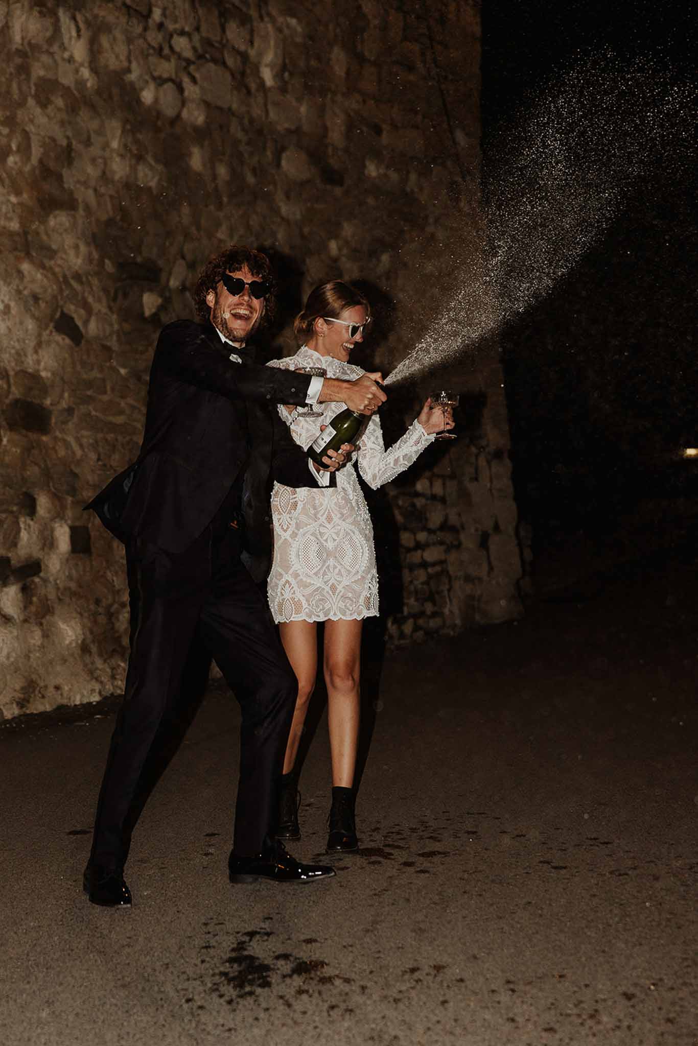 Bride and groom spraying champagne at night against stone wall, wearing sunglasses in rock-and-roll editorial style