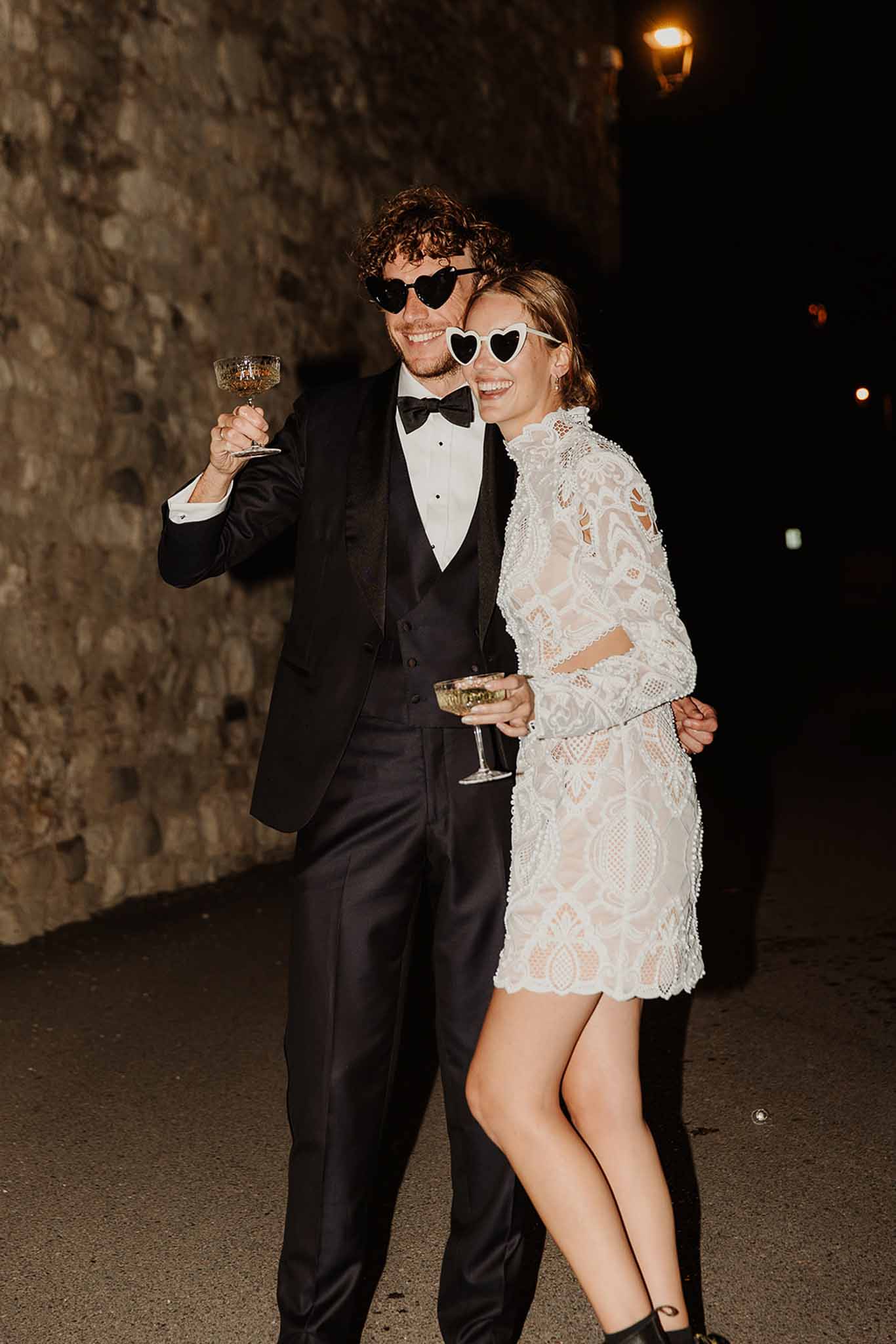 Bride and groom laughing at night holding champagne coupes wearing heart-shaped sunglasses at after party