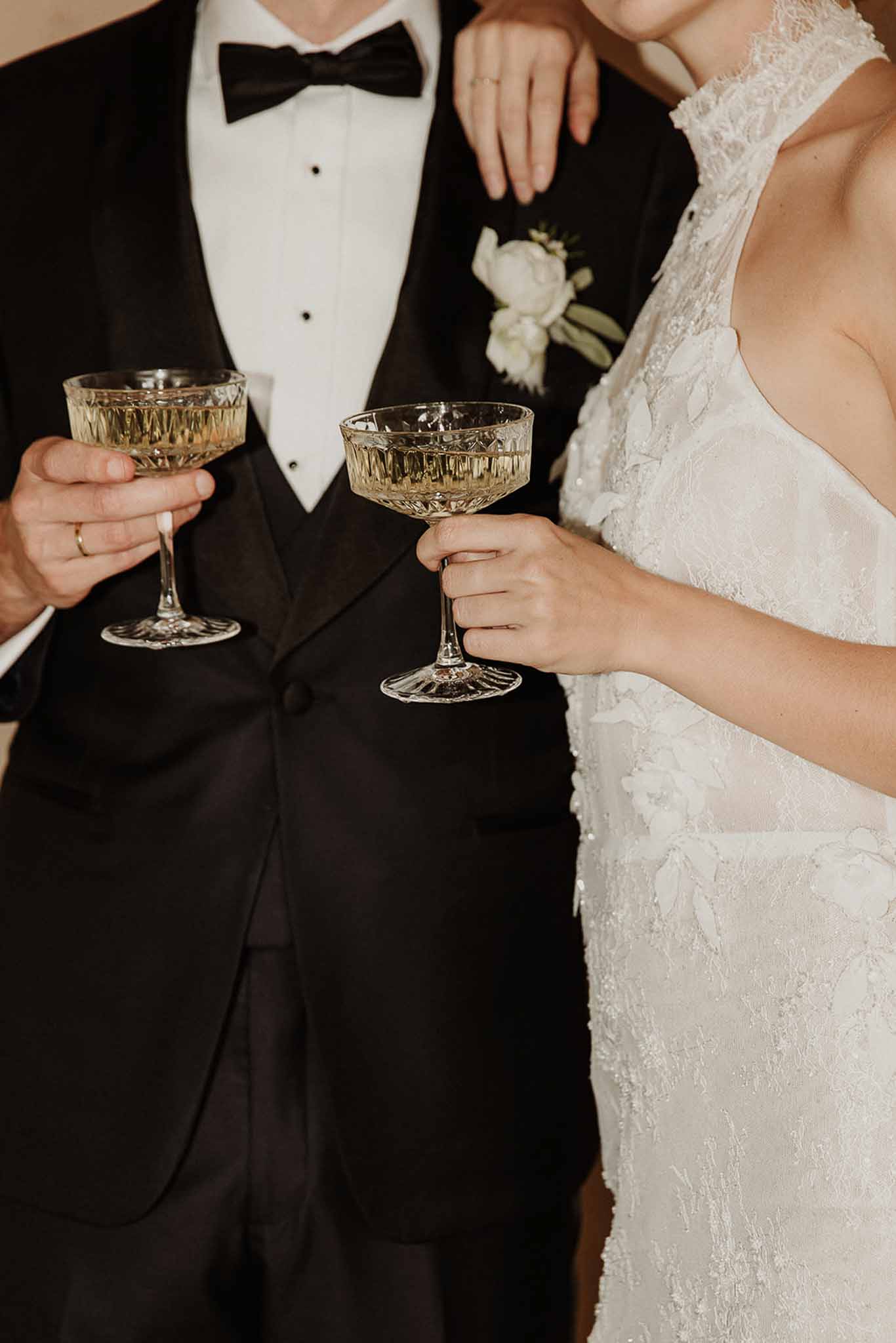 Close-up of bride and groom holding crystal champagne coupes with peony boutonniere and lace gown