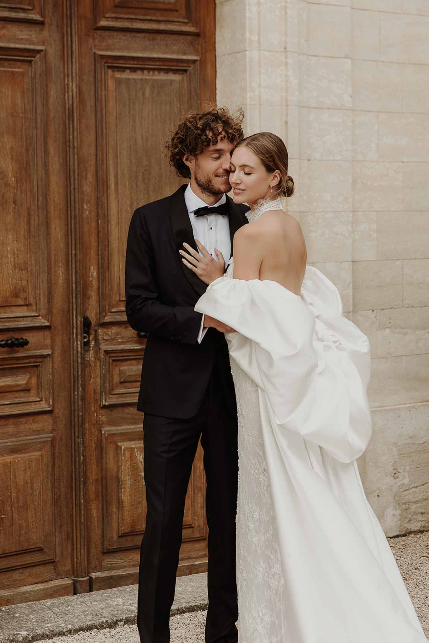 Couple in intimate embrace against chateau wooden doors, bride wearing white structured cape