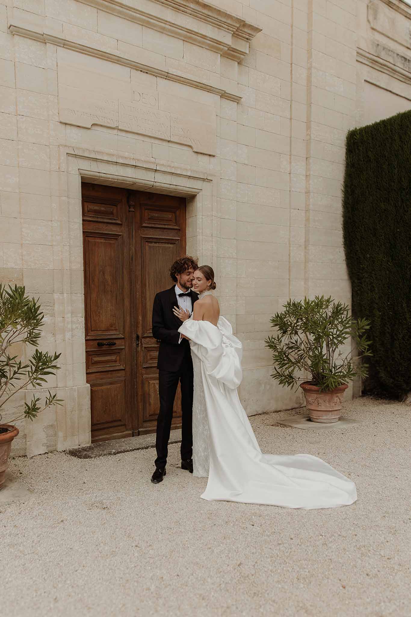 Groom kissing brides temple before chateau chapel doors bride in off-shoulder gown with voluminous puff sleeves and long t...