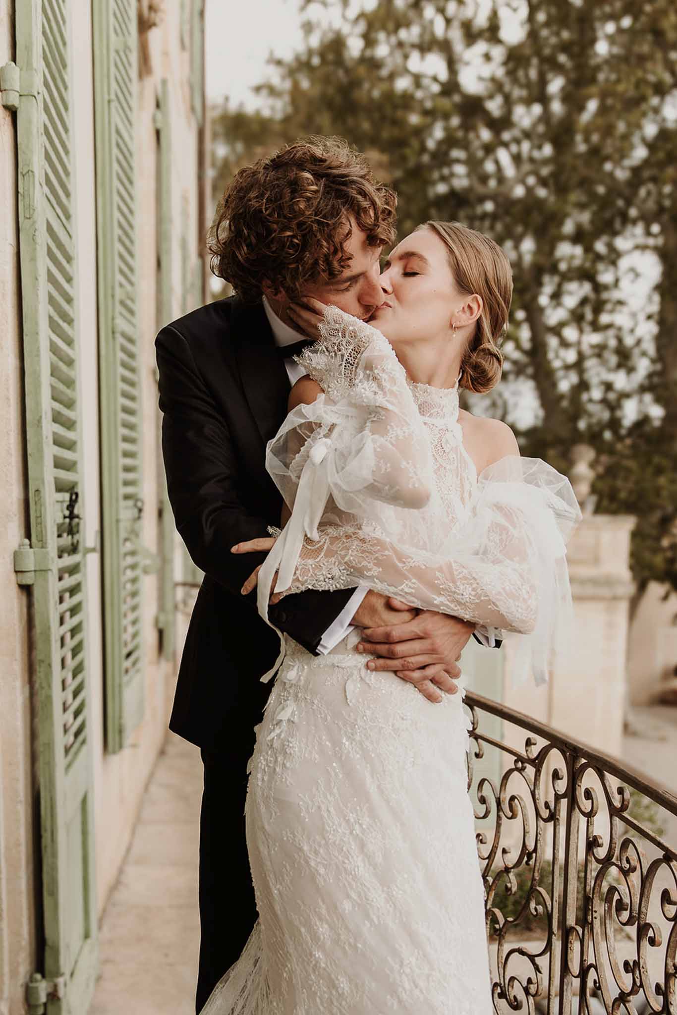 Bride and groom kissing on chateau balcony framed by sage green shutters and wrought iron railing
