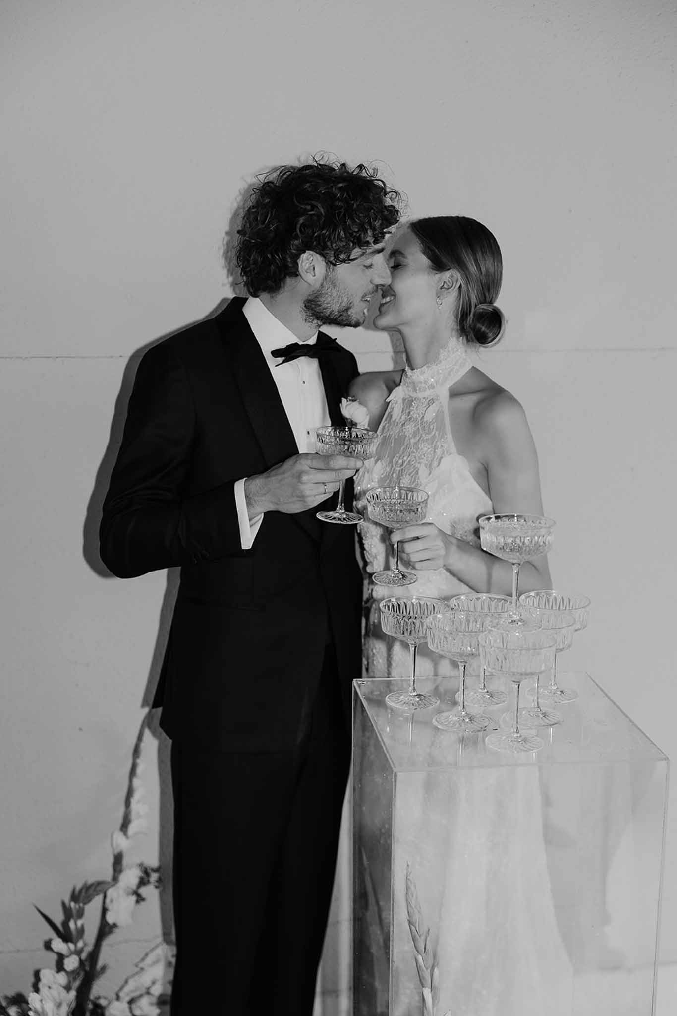 Bride in halter lace gown and groom in tuxedo kiss beside crystal champagne tower in B&W