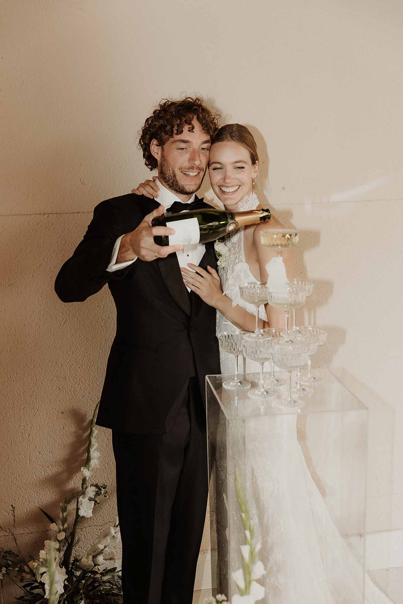 Bride and groom pouring champagne into a crystal coupe tower on an acrylic plinth with white gladiolus at the base